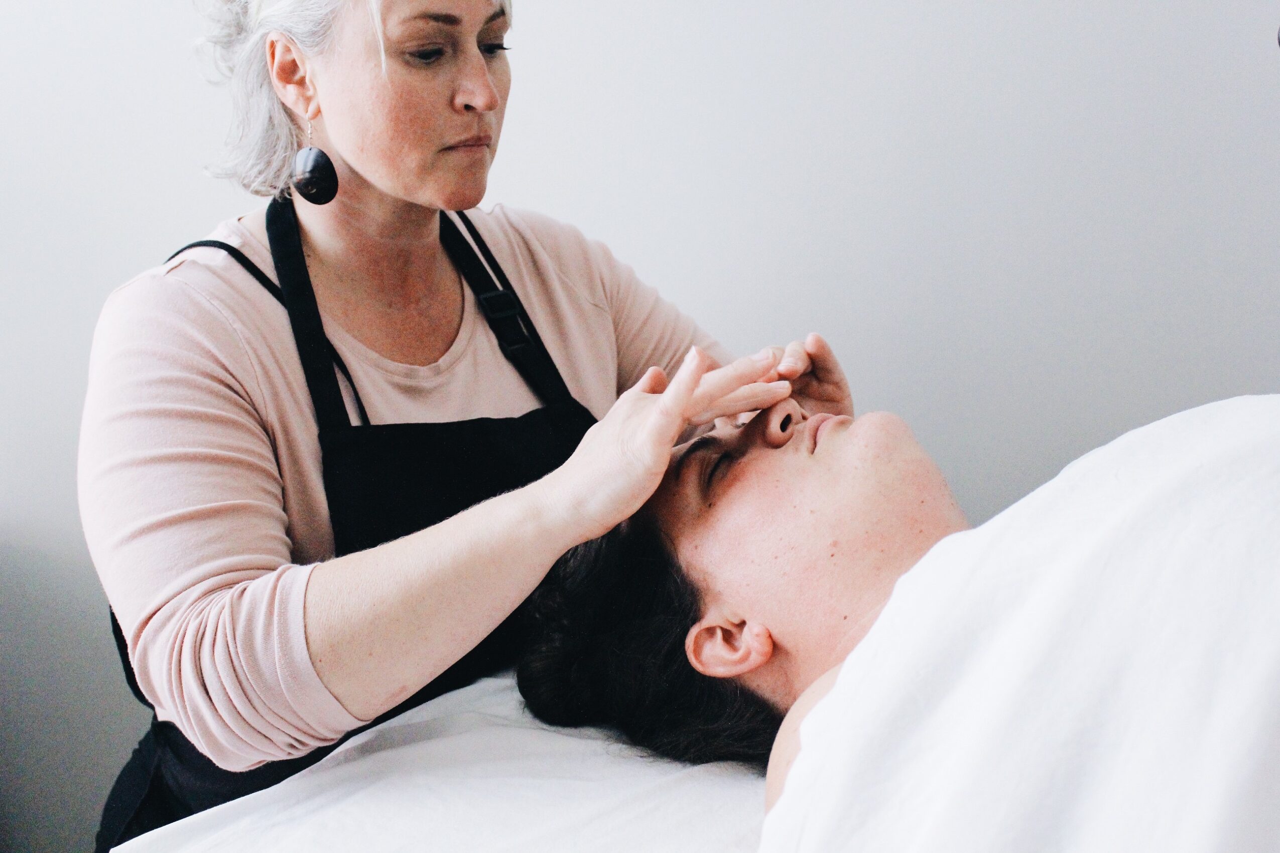 A woman in a black apron gently touches a person's forehead while they lie in bed.