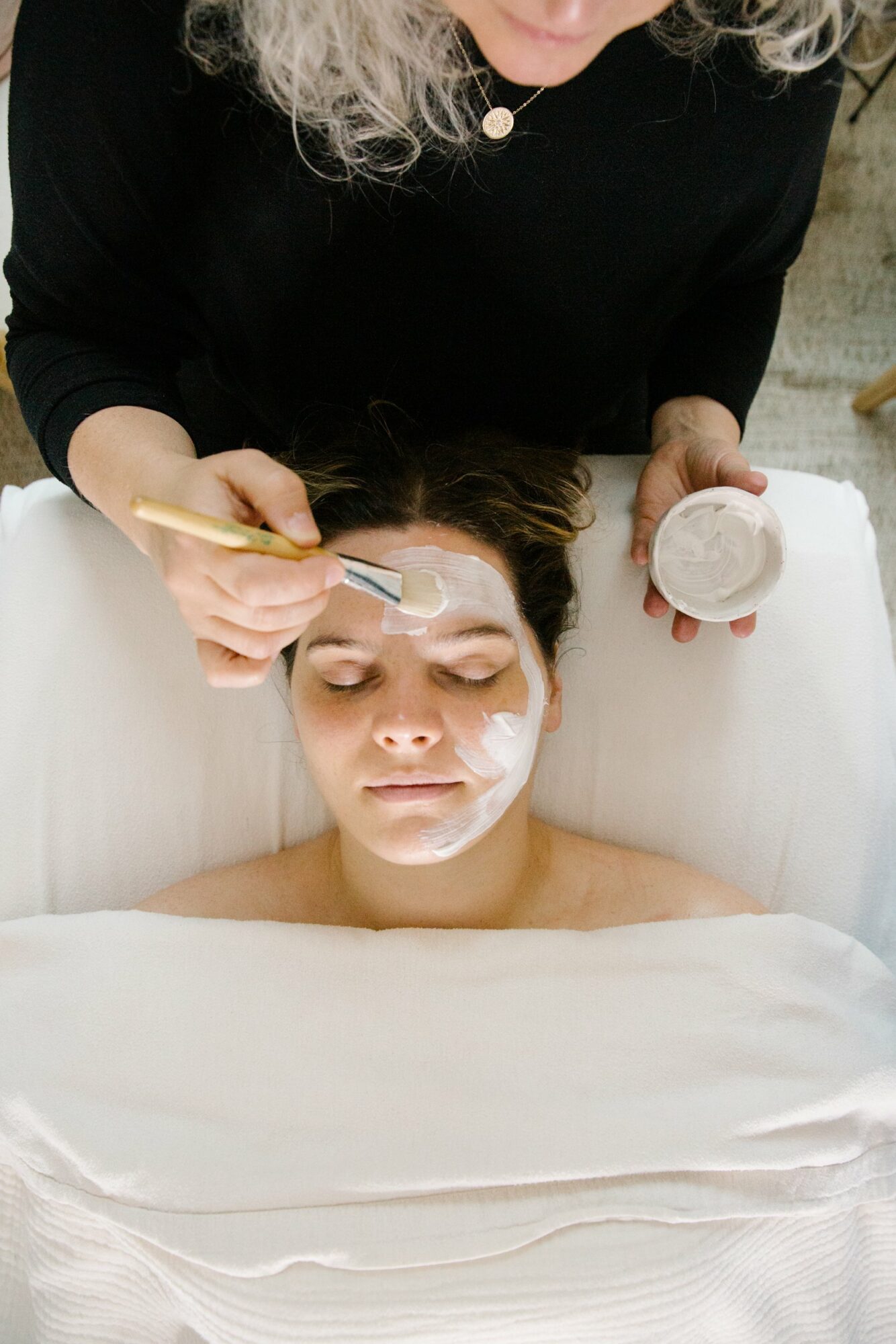 A woman applies facial cream to a young person's face during a skincare treatment, with the person lying down and eyes closed.