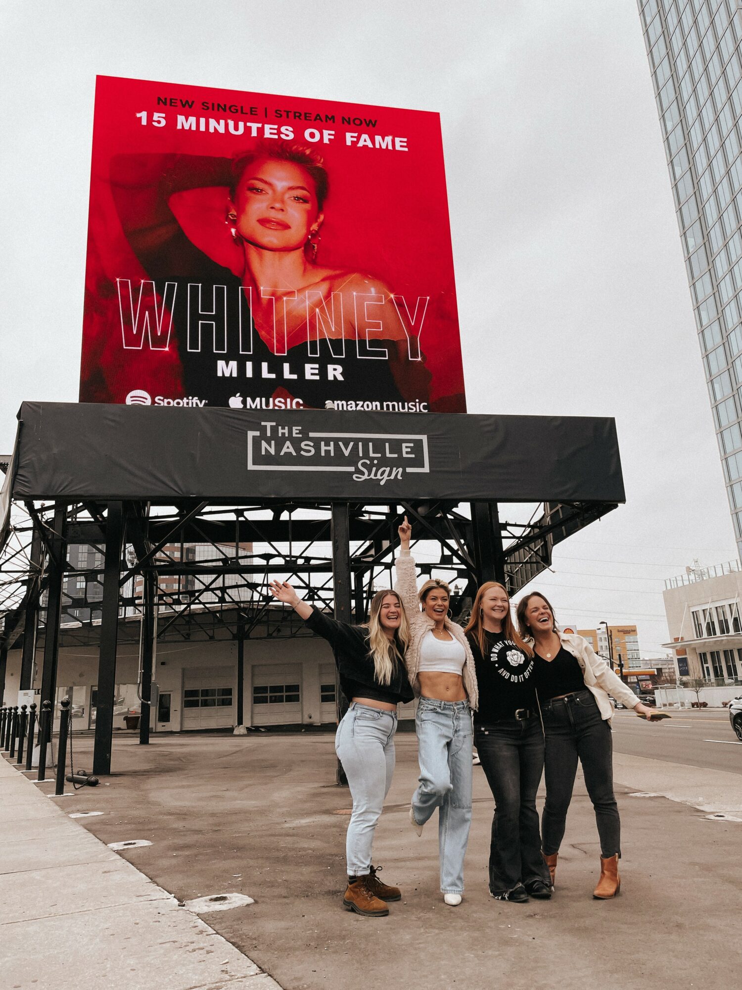 Four women stand together outdoors in front of a black sign with white text, with a large red billboard above them showing a woman and text. Alt text word count: 27. Confidence: 0.9