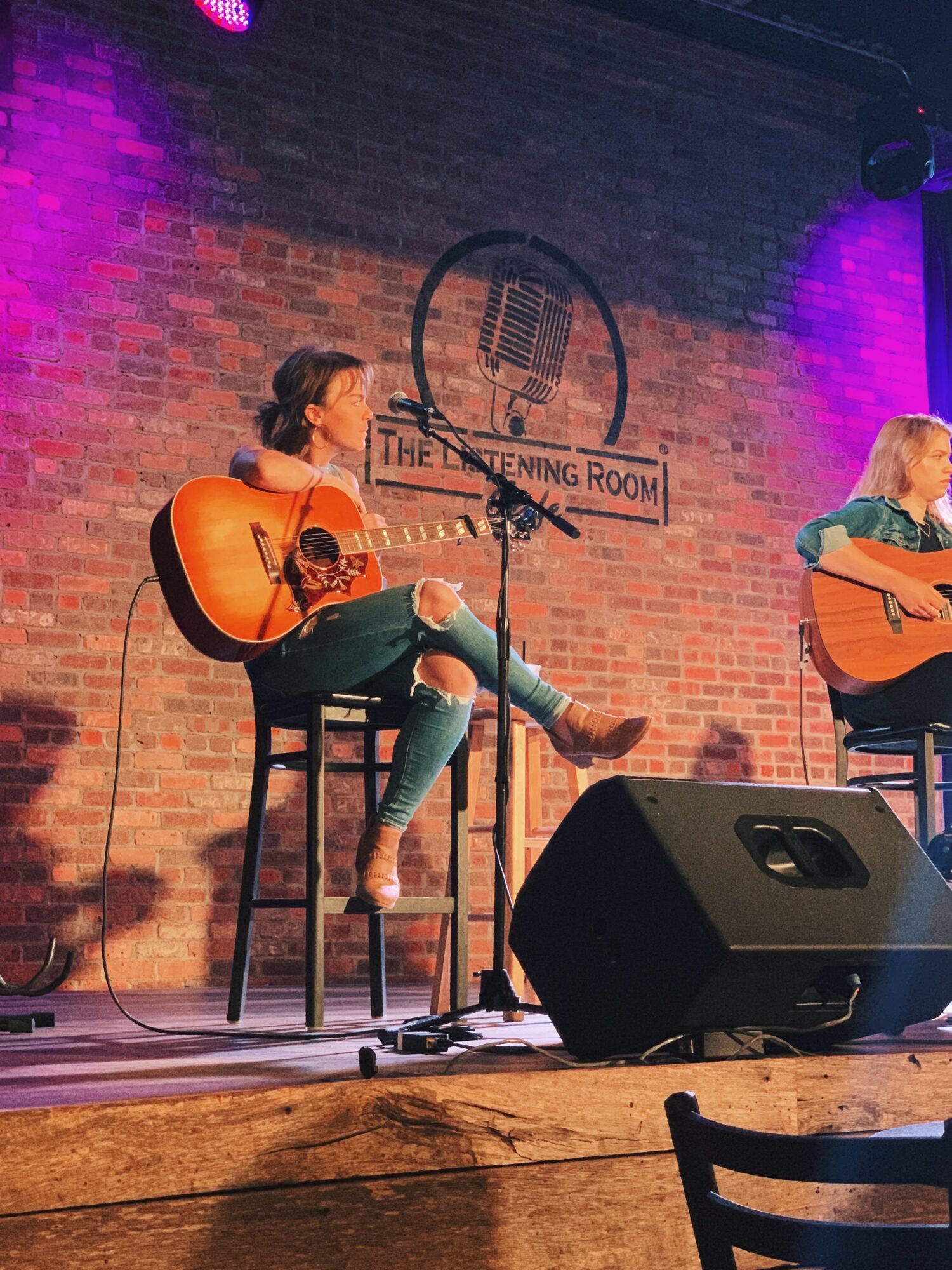 Two women playing guitars on stage in a performance room with a brick wall background and a logo reading 'The Opening Room'.
