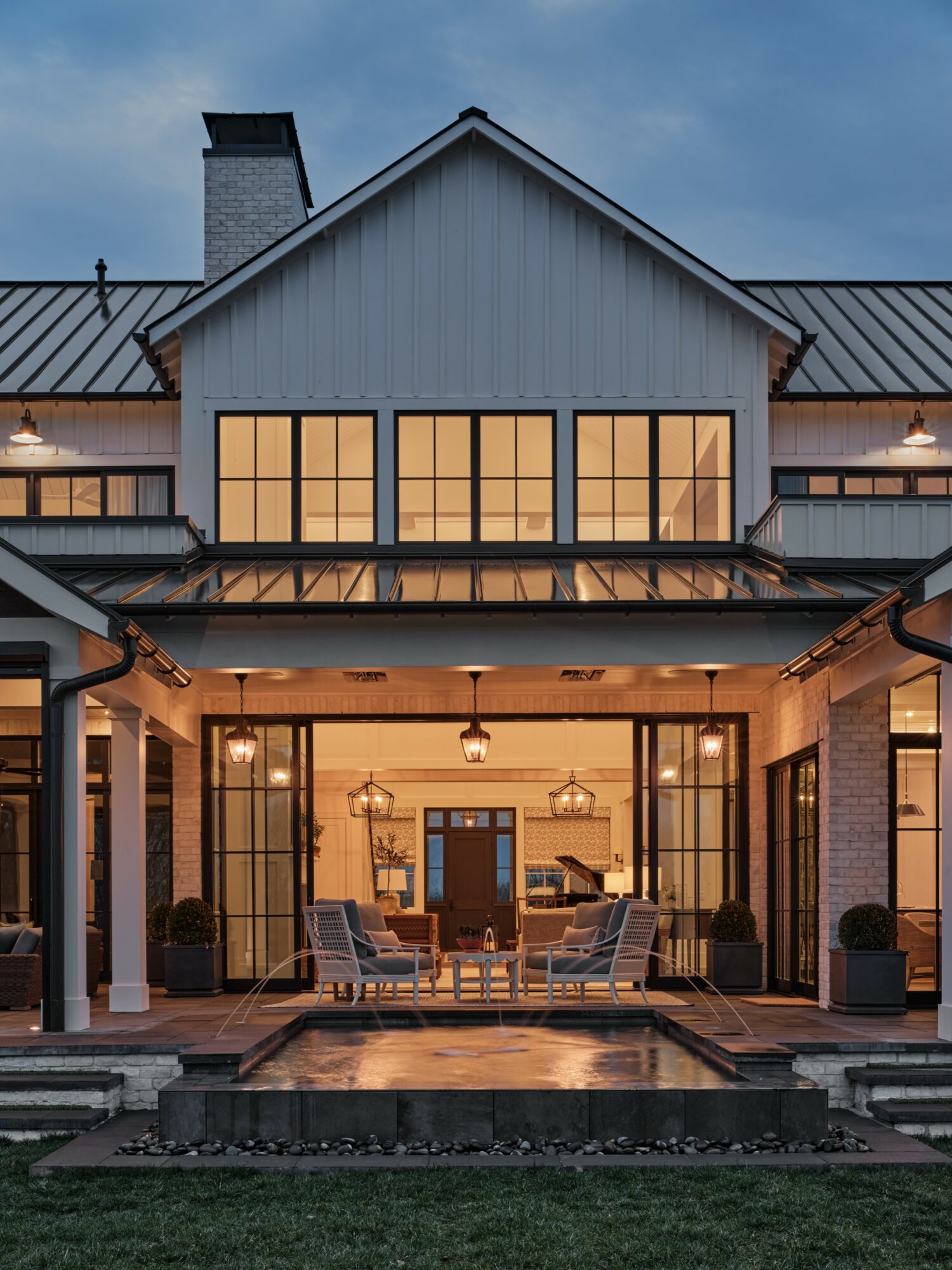 Exterior view of a house with large windows and a covered patio, illuminated at dusk, with a lawn in front.
