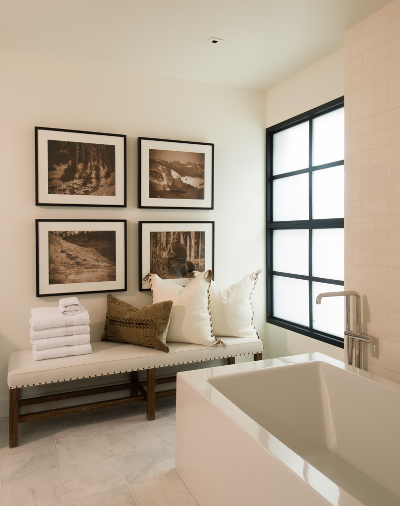 Corner of a room with a window, framed pictures on the wall, a bench with pillows and towels, and a bathtub.