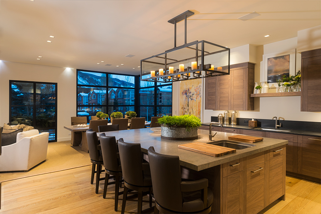 Modern kitchen and dining area with large windows, a chandelier above a wooden island, and seating for six, with a living space in the background.