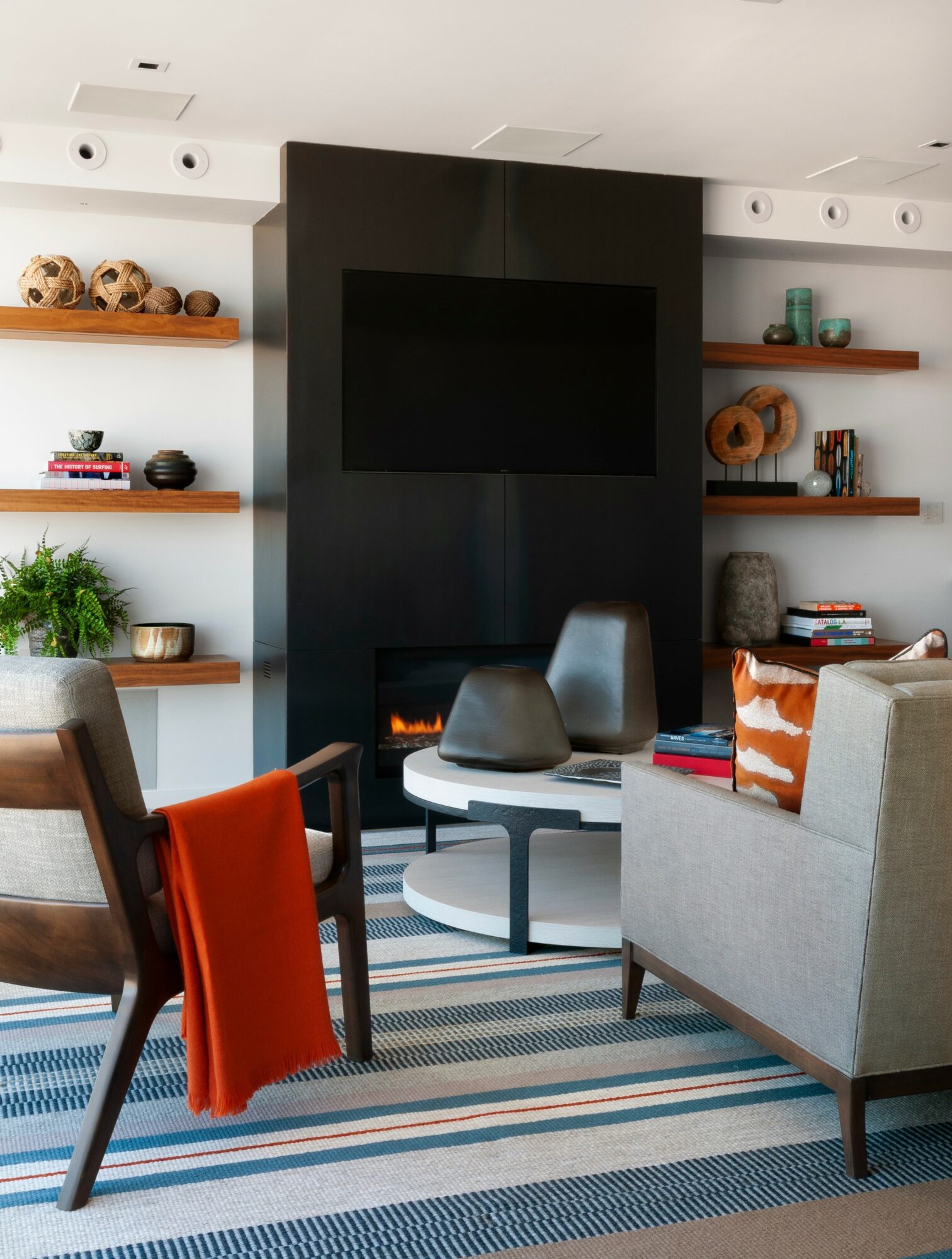 Living room with a black fireplace, wall-mounted TV, shelves with decor, and seating including a gray armchair and a wooden chair with an orange blanket.