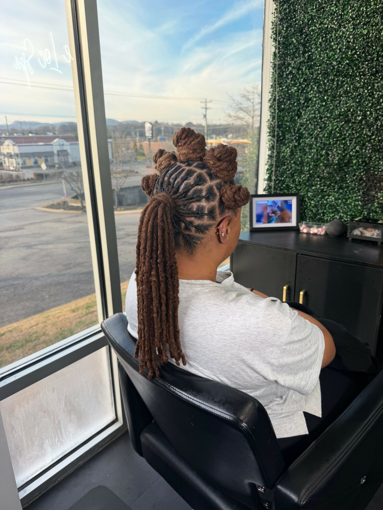 Person with long braided hair sitting in a chair, looking out a window at a cityscape and sky.