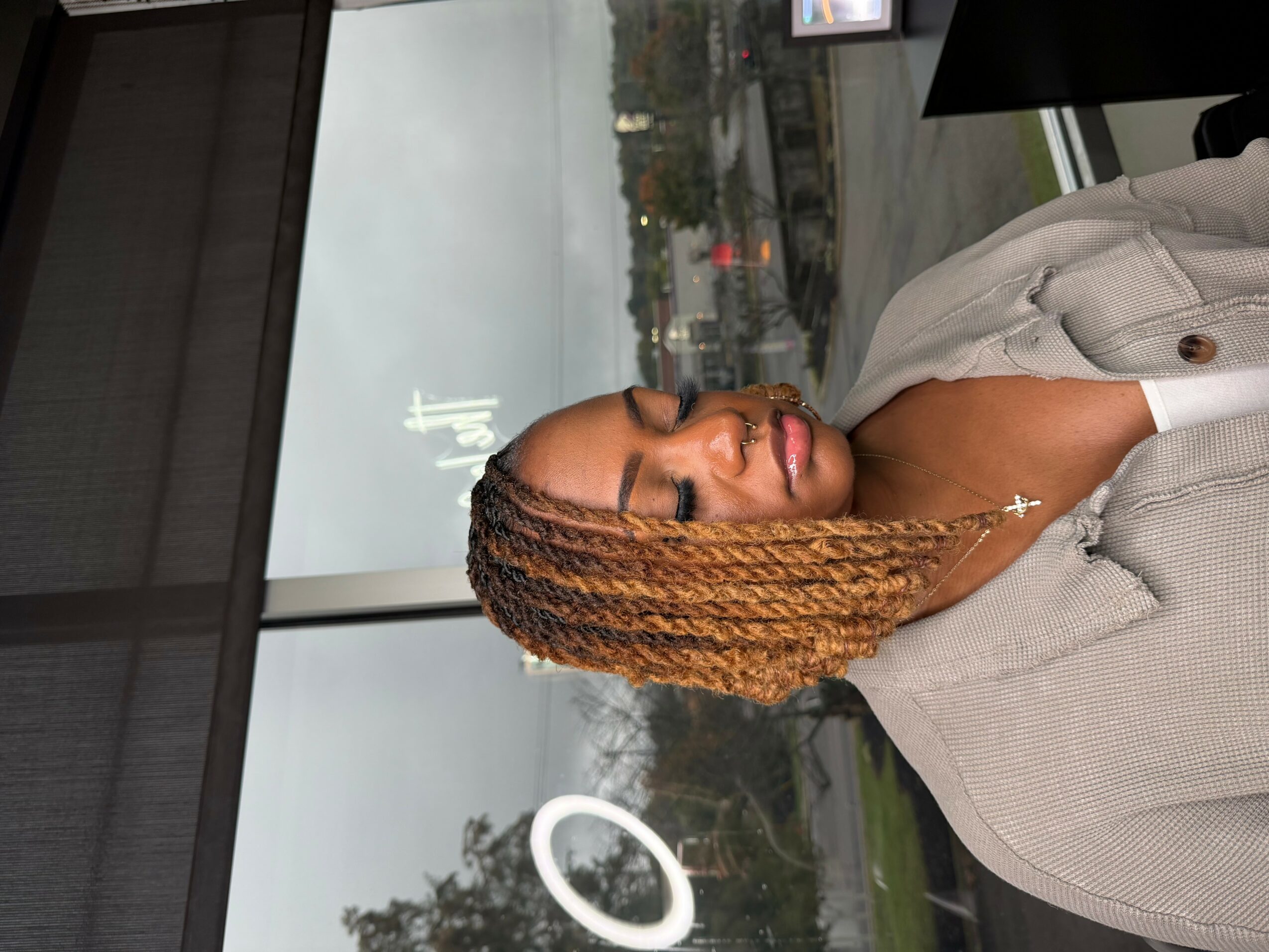 Woman with braided hair and a cross necklace, looking out a window on a cloudy day.