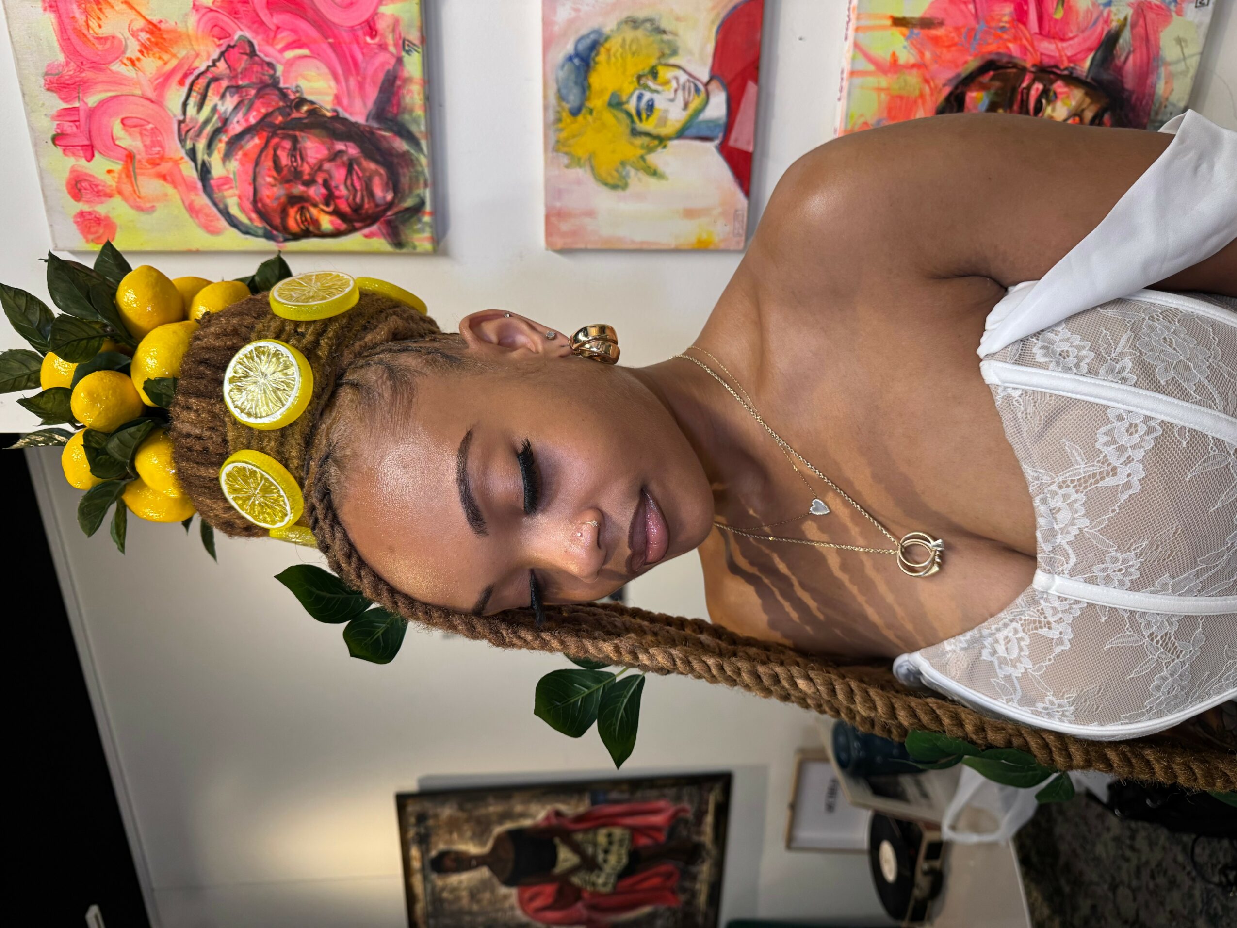 Woman with braided hair, lemon headpiece, and white lace top, smiling with eyes closed, in front of colorful paintings.