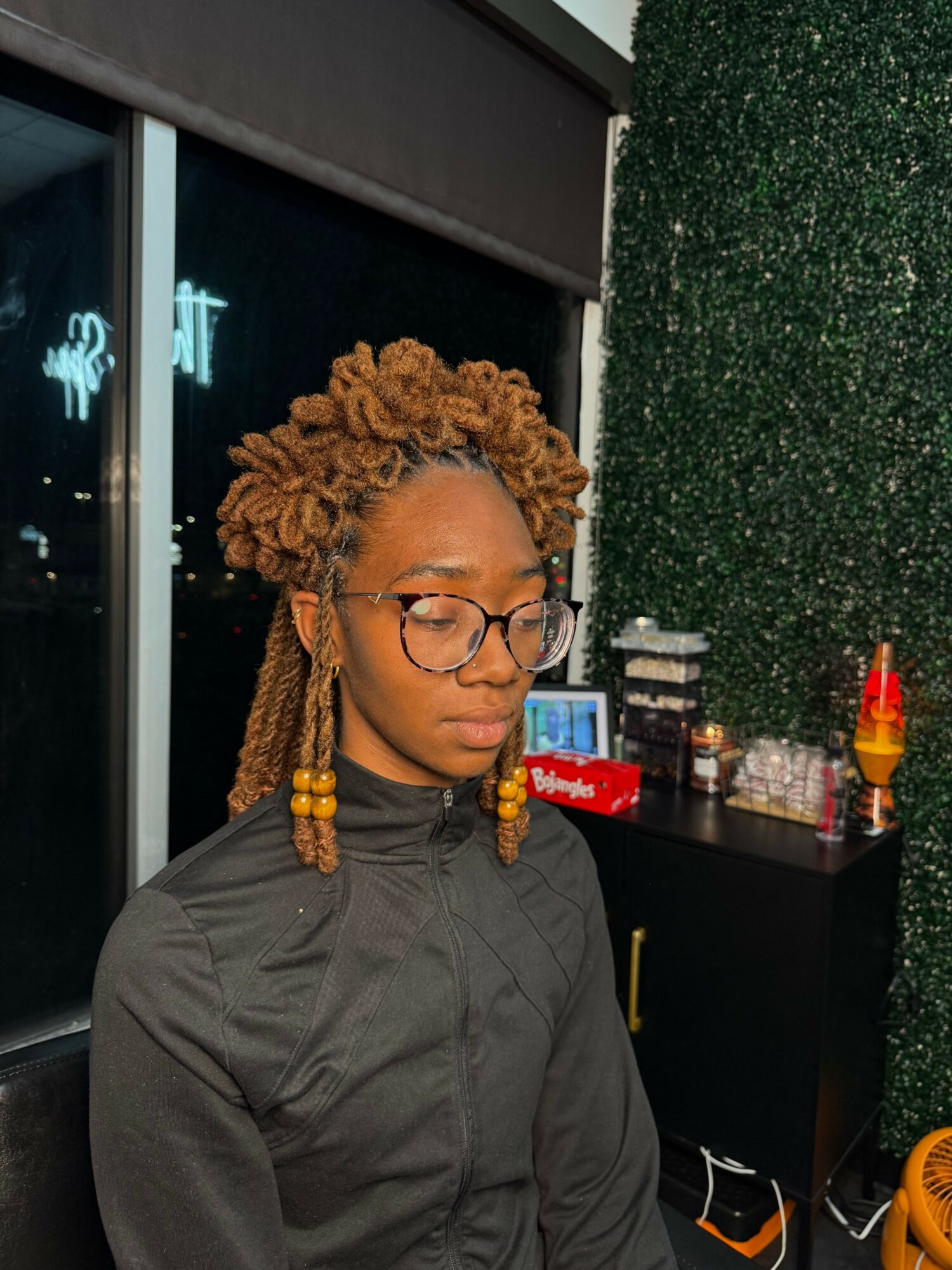 Person with curly hair and glasses smiling, standing indoors near a window with a green wall behind.