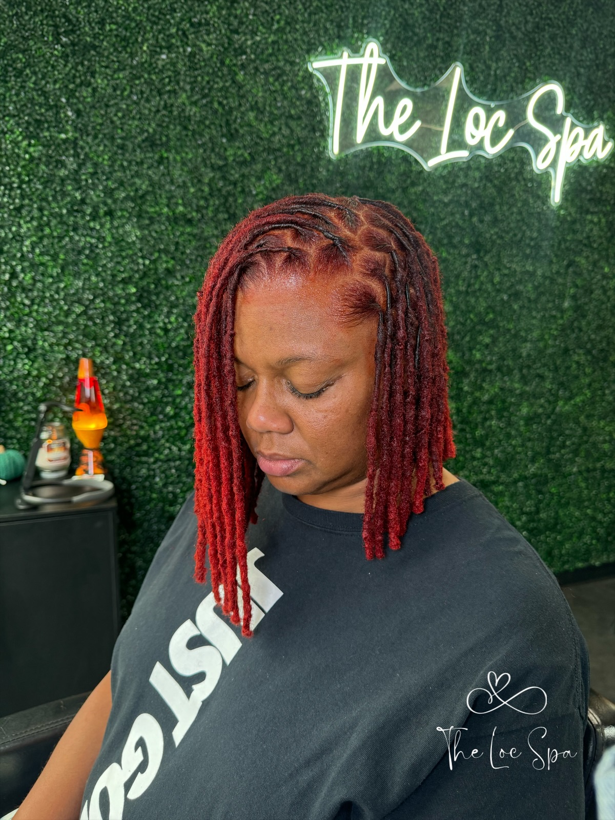 Woman with red dreadlocks looking down, standing in front of a green wall with neon sign reading 'The Loc Spa'.
