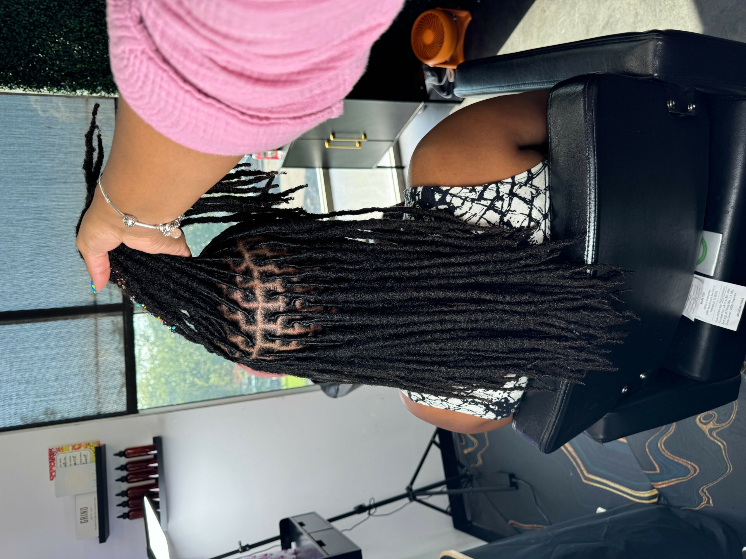 Person with long dreadlocks sitting in a chair, wearing a black and white patterned dress, in a room with a window and shelves.