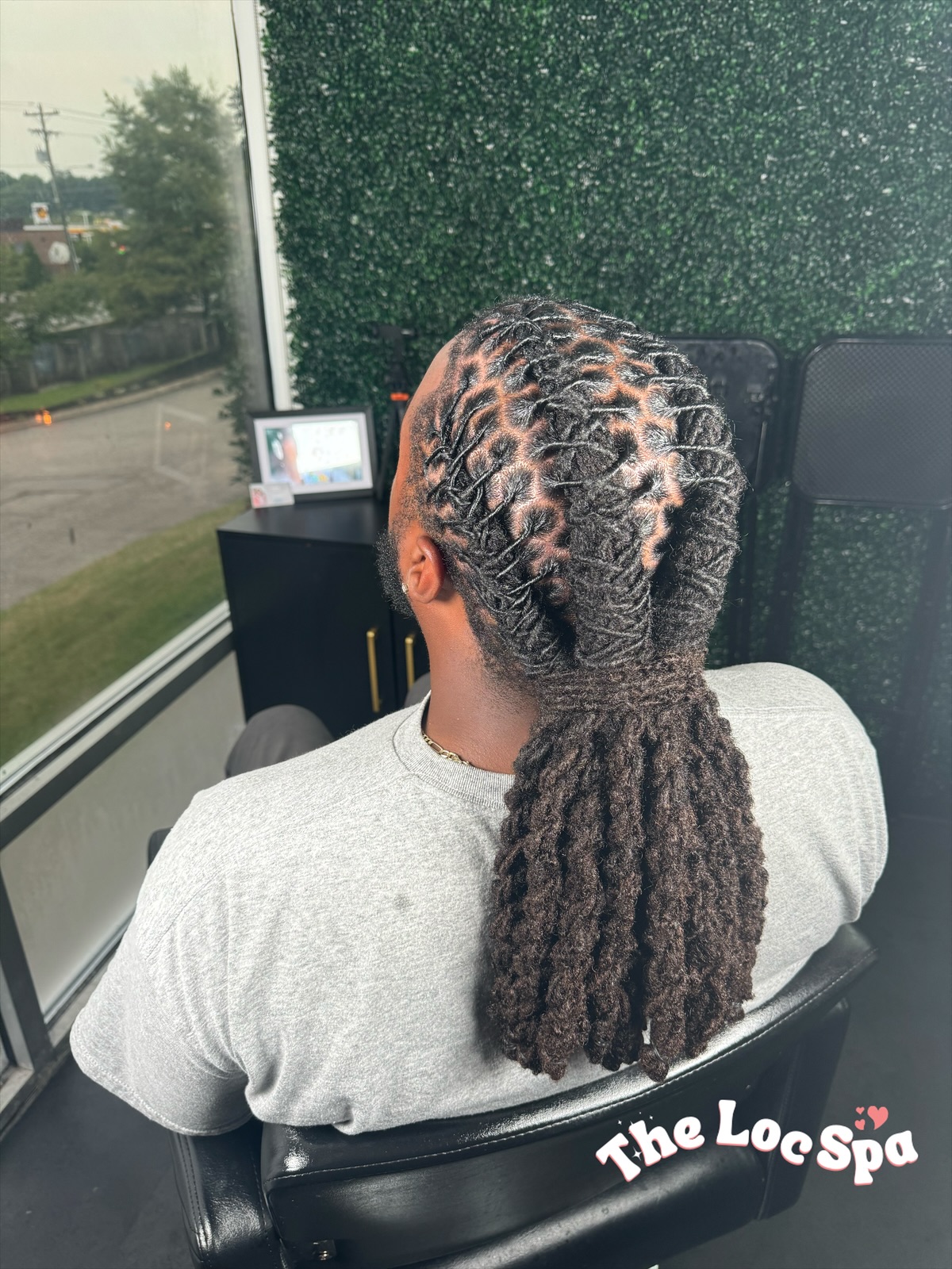 Person with styled braids and dreadlocks sitting in a salon chair, facing away, near a window with a green wall background.