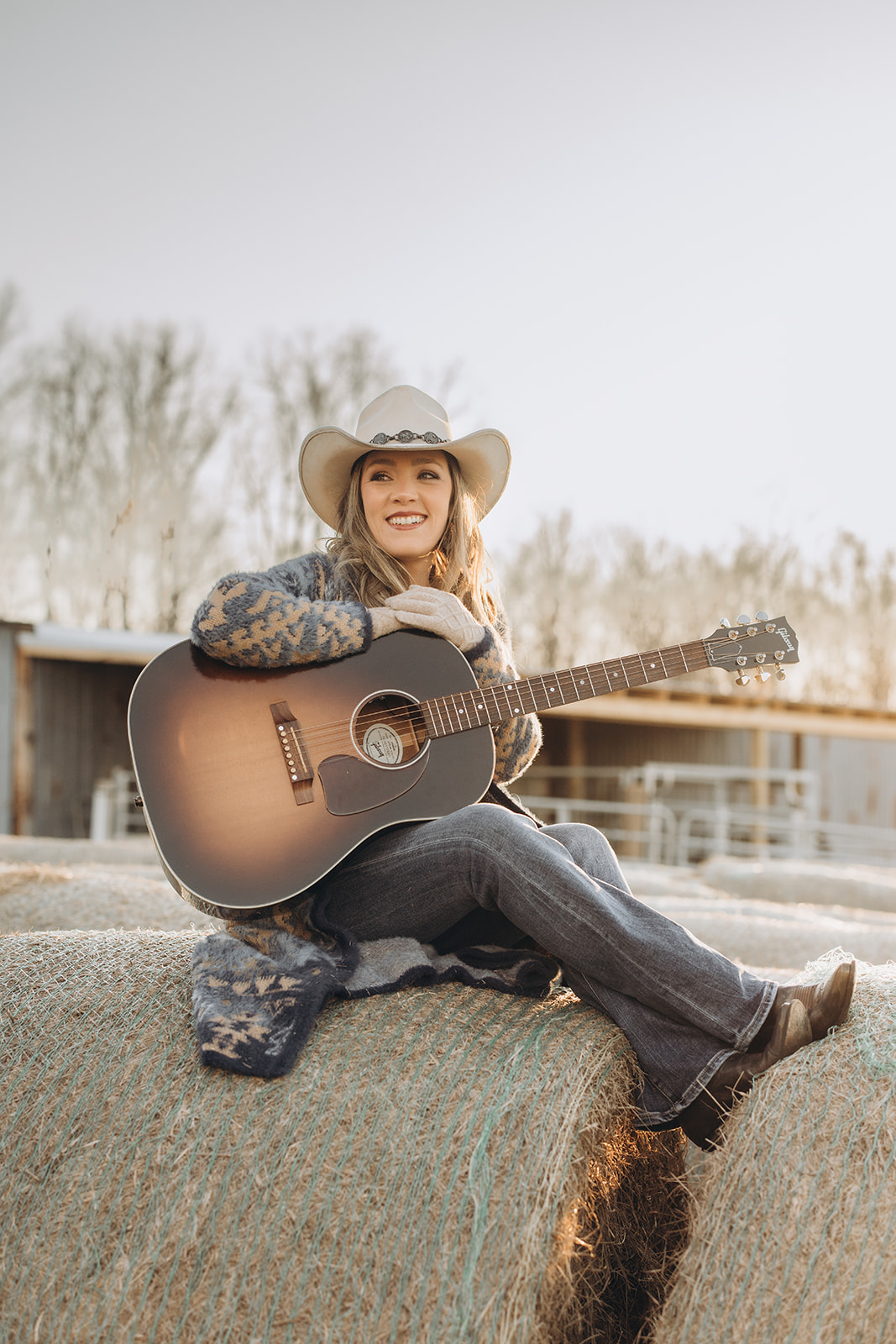 Woman wearing a cowboy hat sitting outdoors with a guitar, smiling, with trees and a fence in the background.