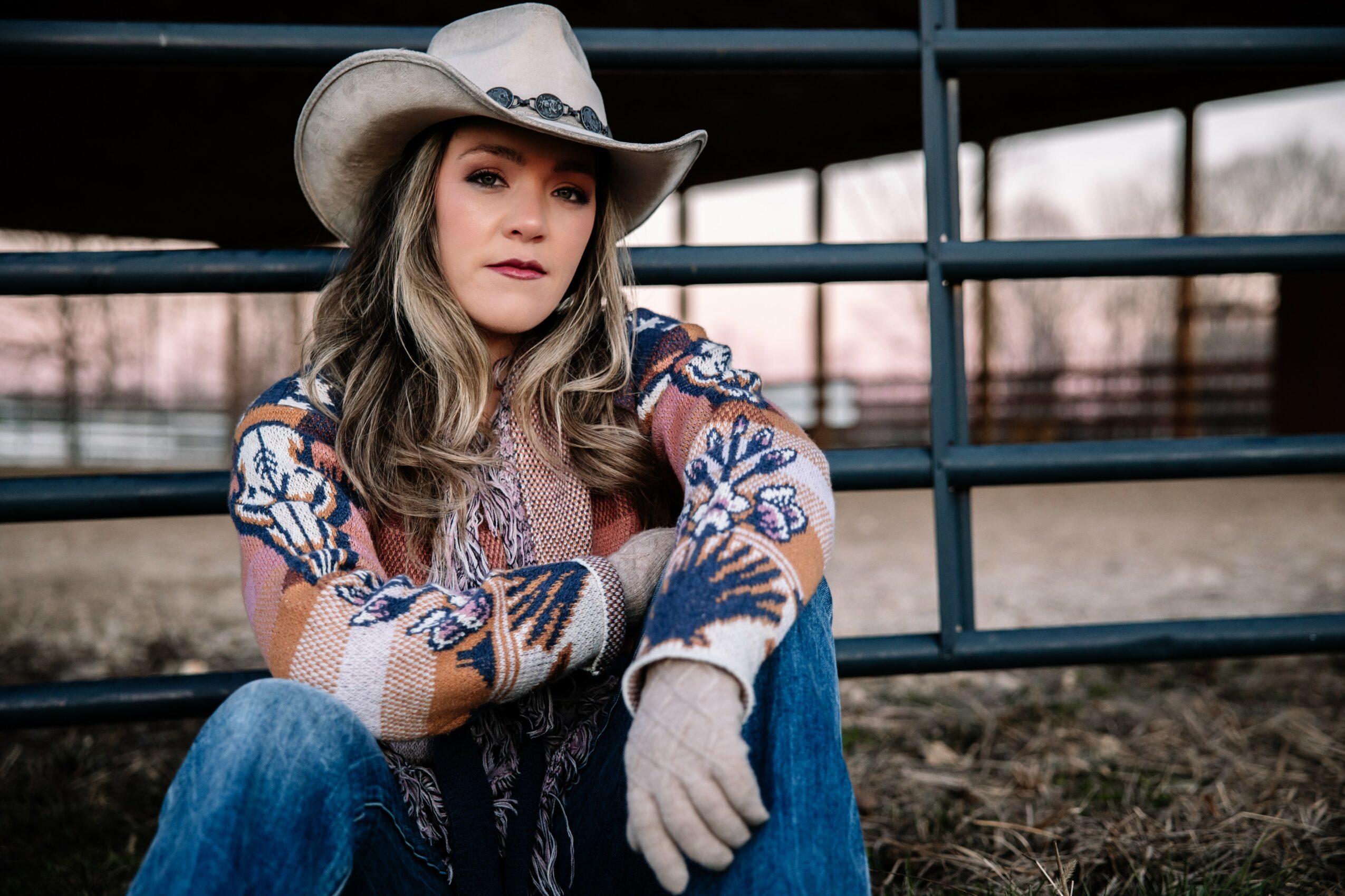 Woman wearing a cowboy hat and patterned sweater, sitting outdoors near a metal fence, looking at the camera.