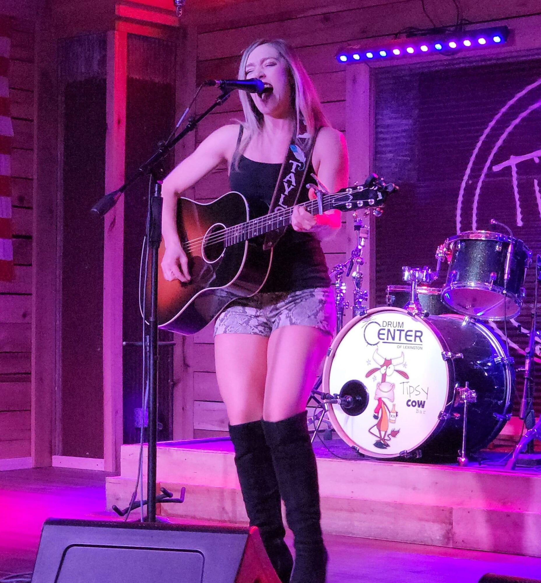 Young woman singing and playing guitar on stage with drum set in background, purple lighting, wooden backdrop, and microphone stand