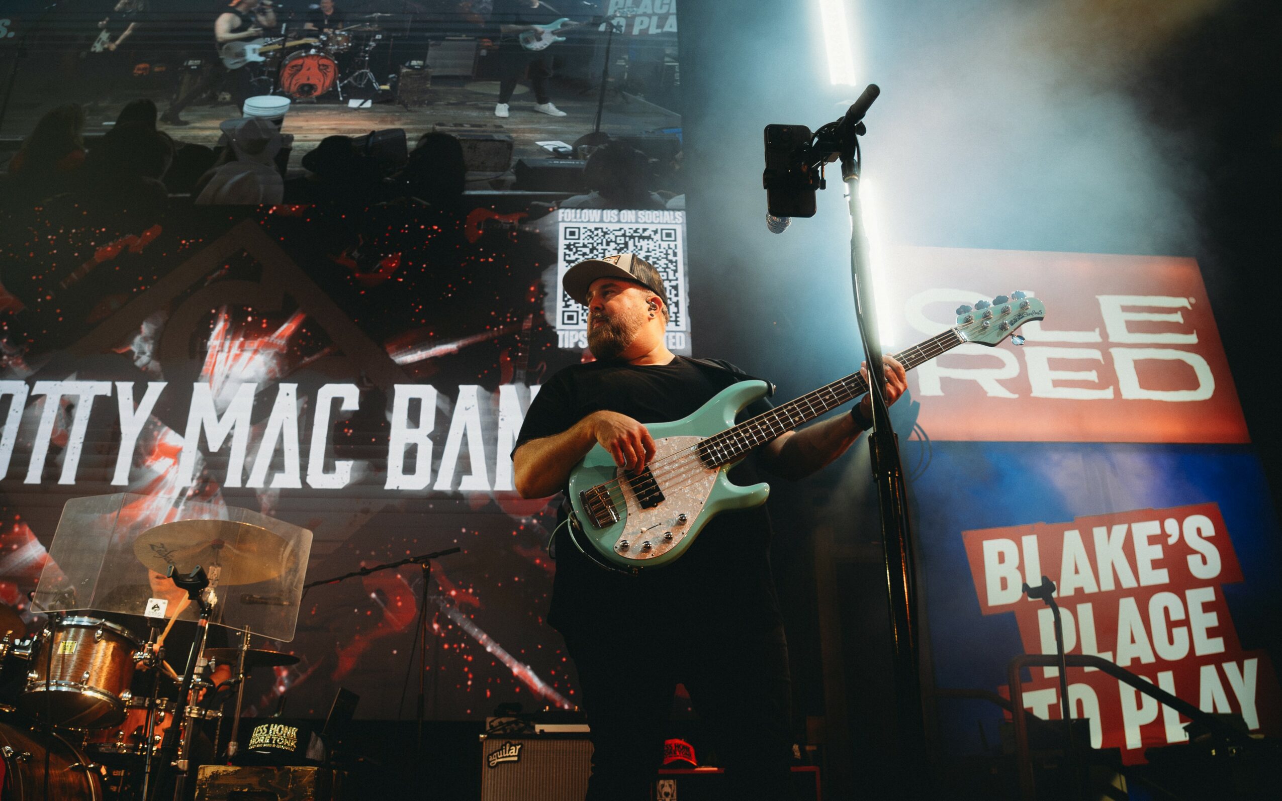 Musician playing bass guitar on stage with a large screen behind displaying text and images.
