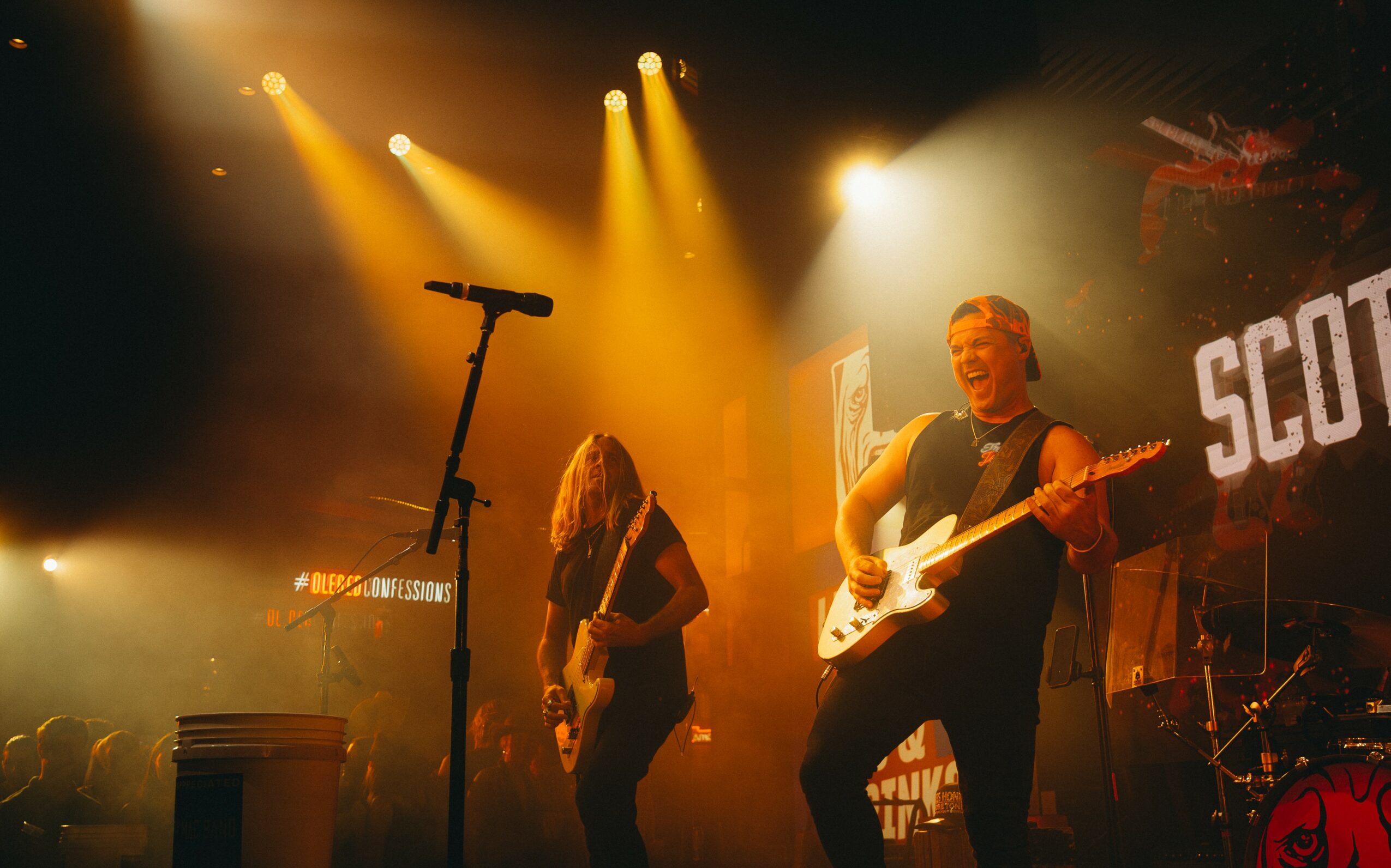 Two musicians perform on stage with bright yellow lights, one playing guitar and the other with long hair, in front of a screen.