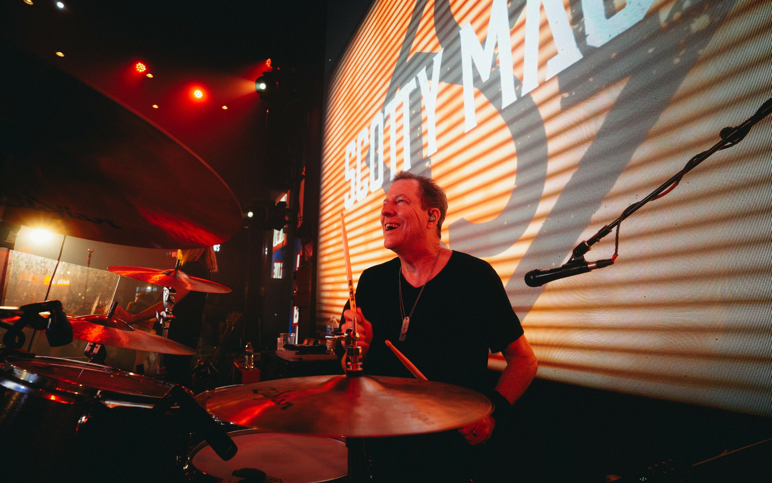 Person playing drums on stage with a large screen displaying text behind them.