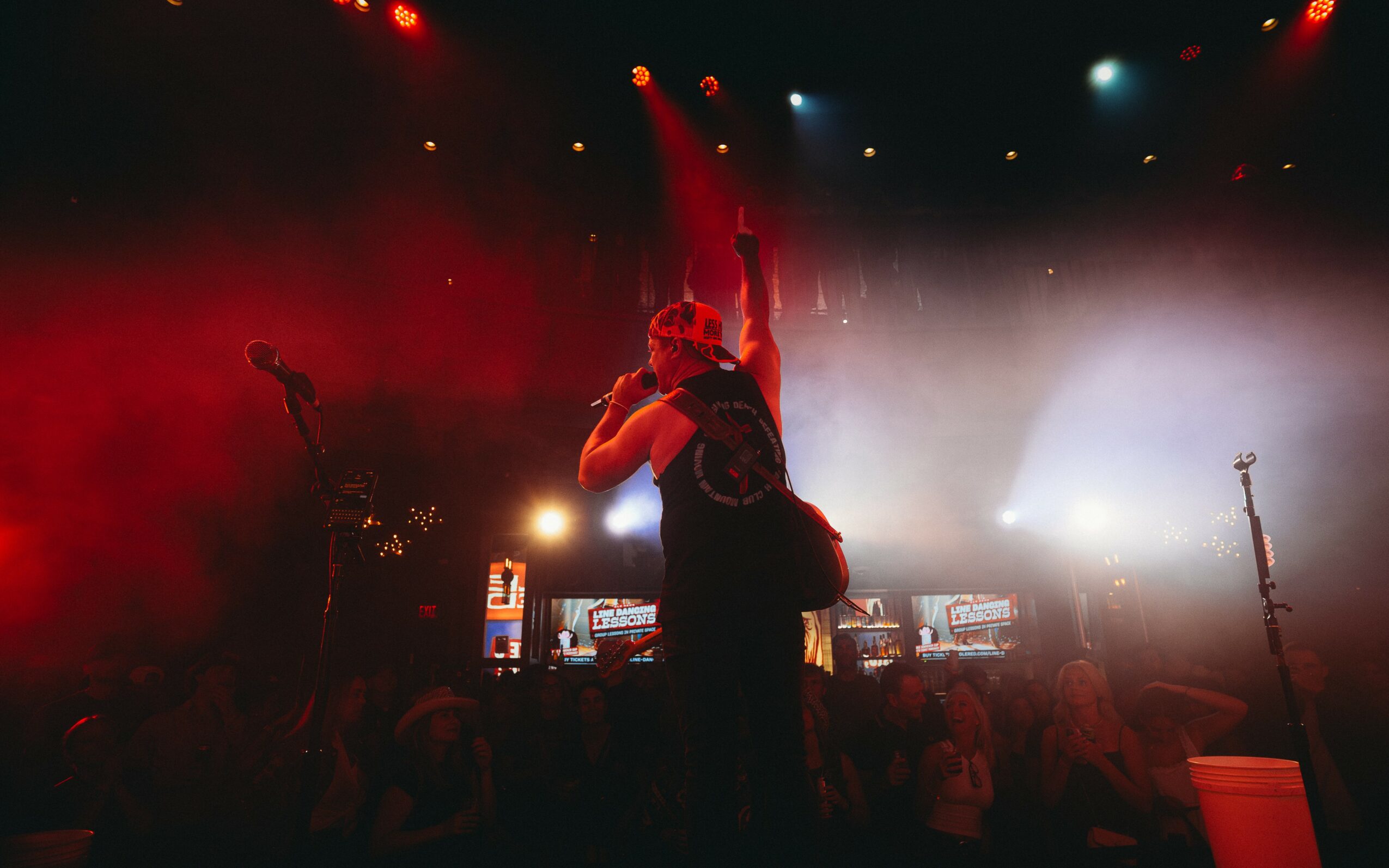 Performer on stage with one arm raised, surrounded by colorful lights and an audience, in a concert setting.
