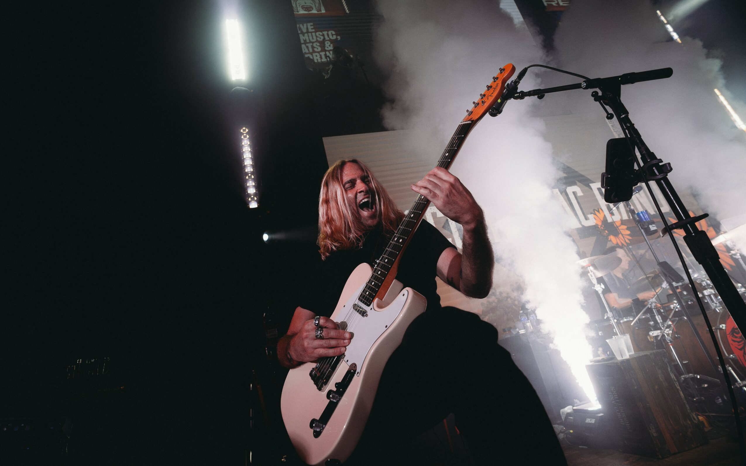 Person playing electric guitar on stage with bright lights and smoke effects, microphone stand nearby.