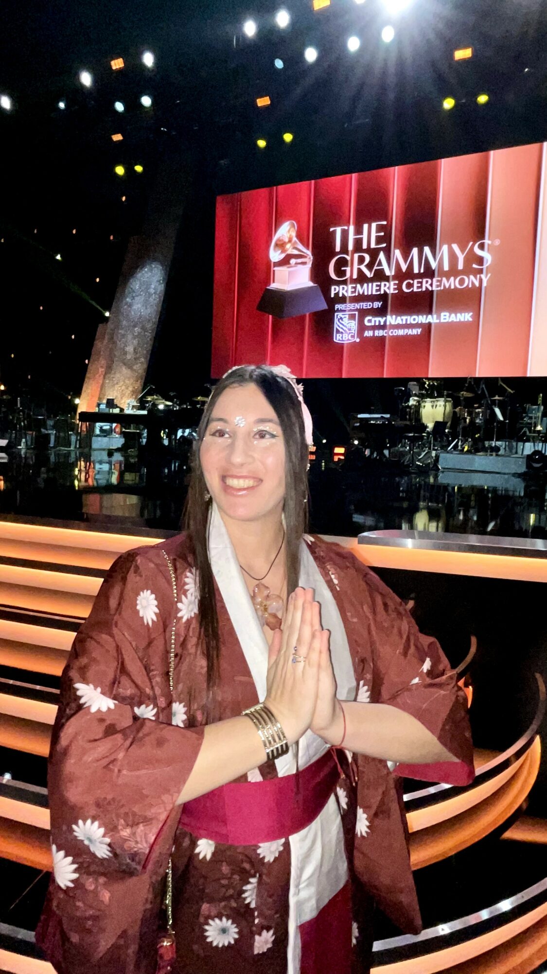 Woman in traditional attire with hands together at an awards ceremony stage, large screen behind displaying 'The Grammys' logo.