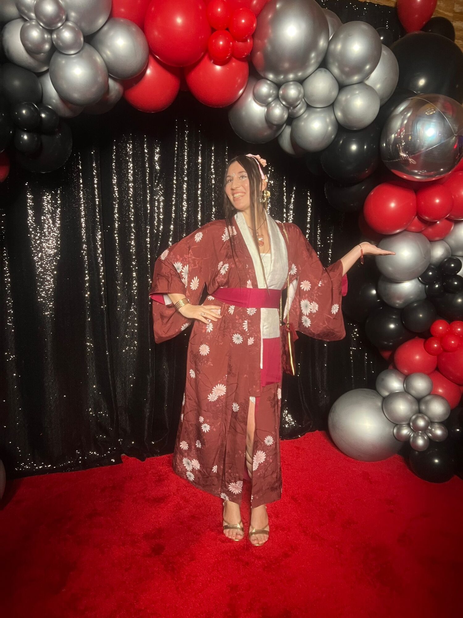 Woman in traditional kimono standing on red carpet with black, red, silver, and gray balloon arch background.