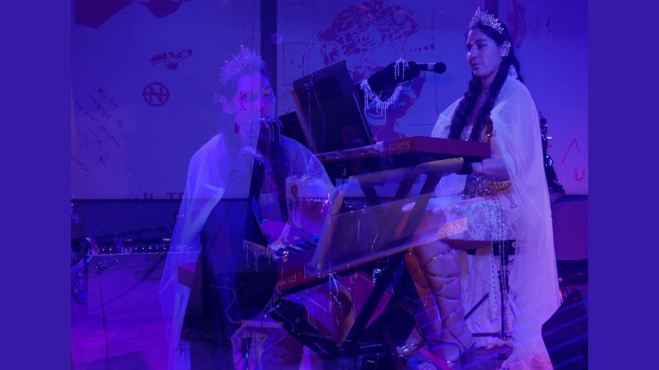Two children dressed in traditional clothing perform on stage with a piano, under purple lighting.