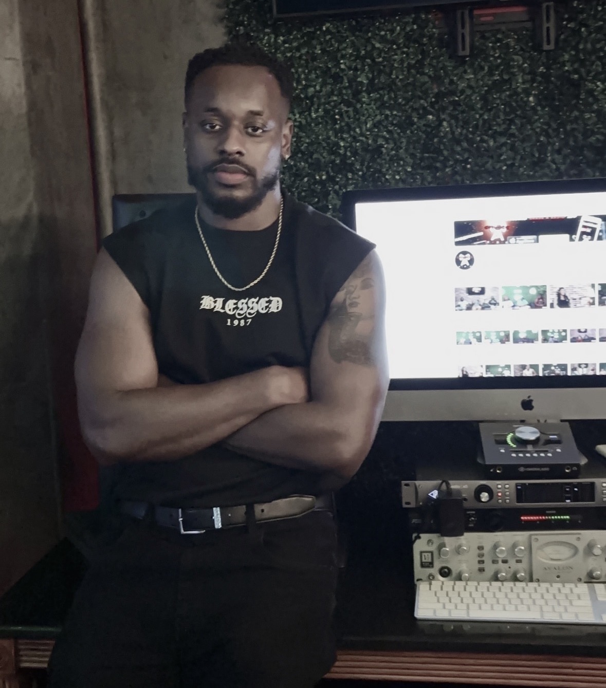Man with crossed arms, wearing a sleeveless shirt, standing in front of a computer monitor and audio equipment, in a room with dark decor.