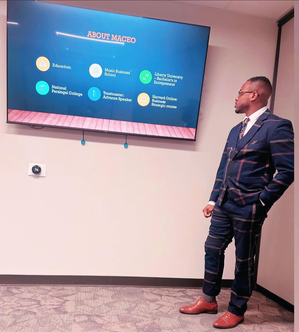 Man in a suit standing in a room with a large screen displaying a presentation slide, facing the screen, with a plain wall and door nearby.
