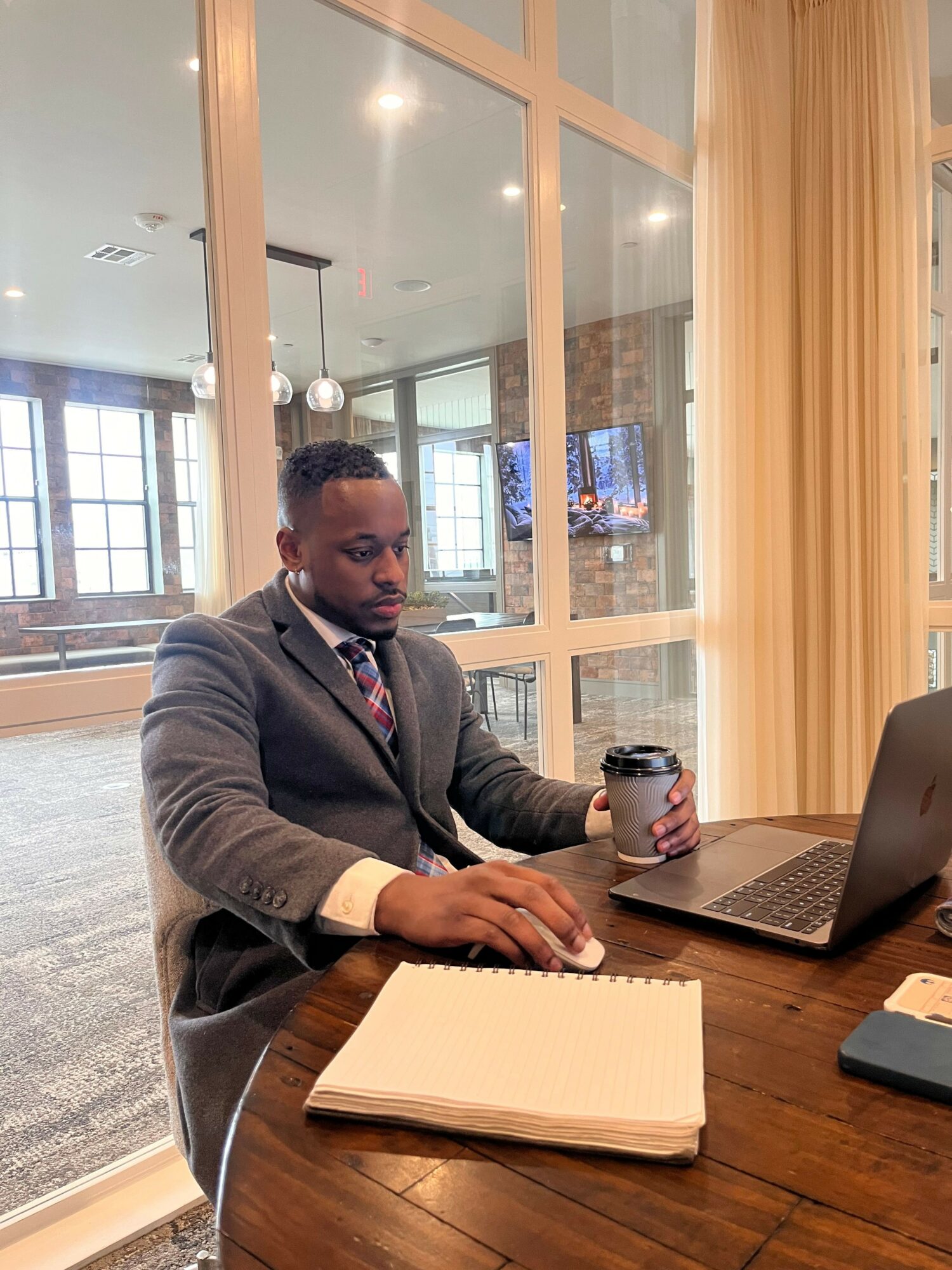 Man in suit sitting at a wooden table with a notebook, laptop, and water bottle in a modern office space.