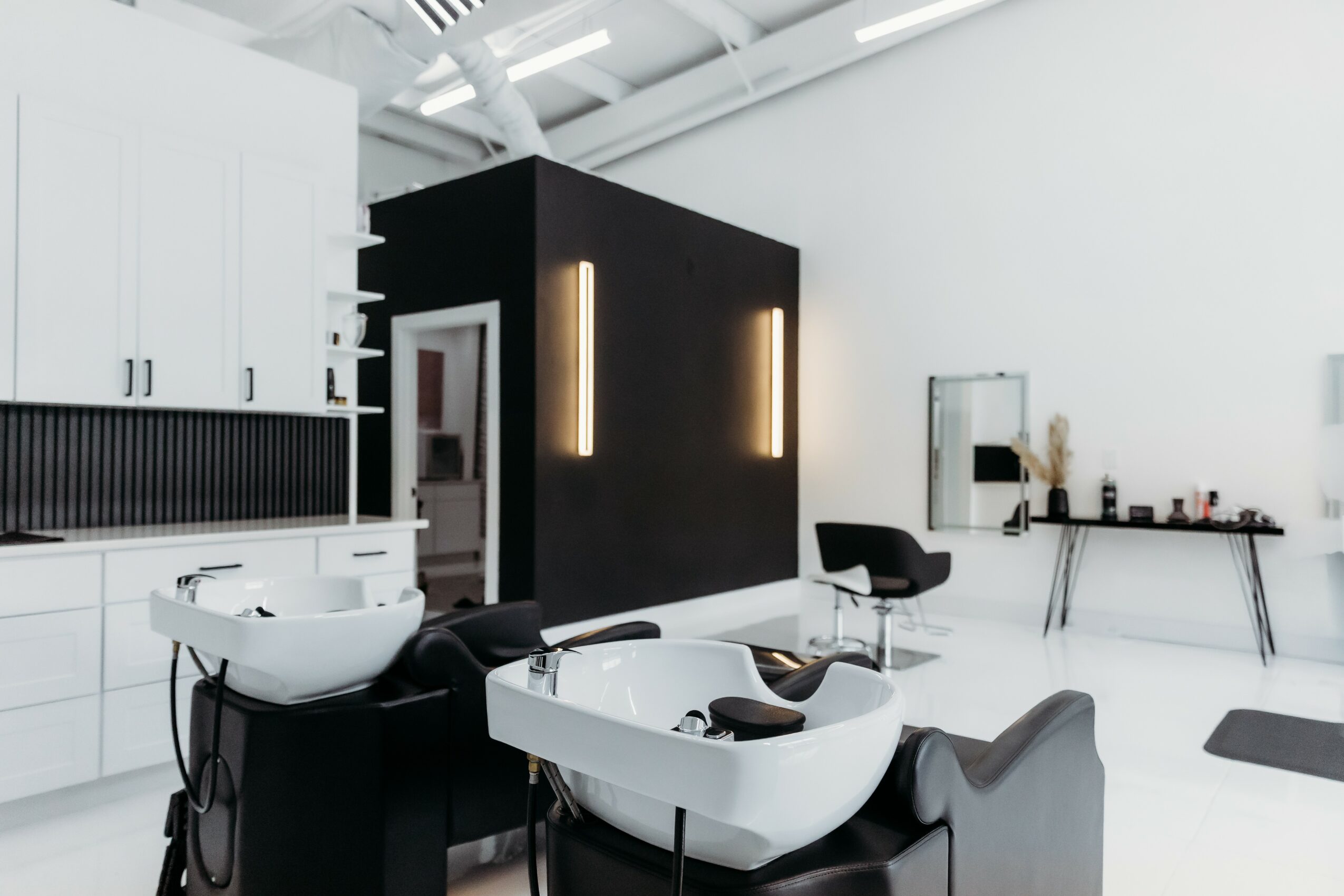 Salon with white sinks, black wall, and minimal decor, including a black chair and a table with beauty products.