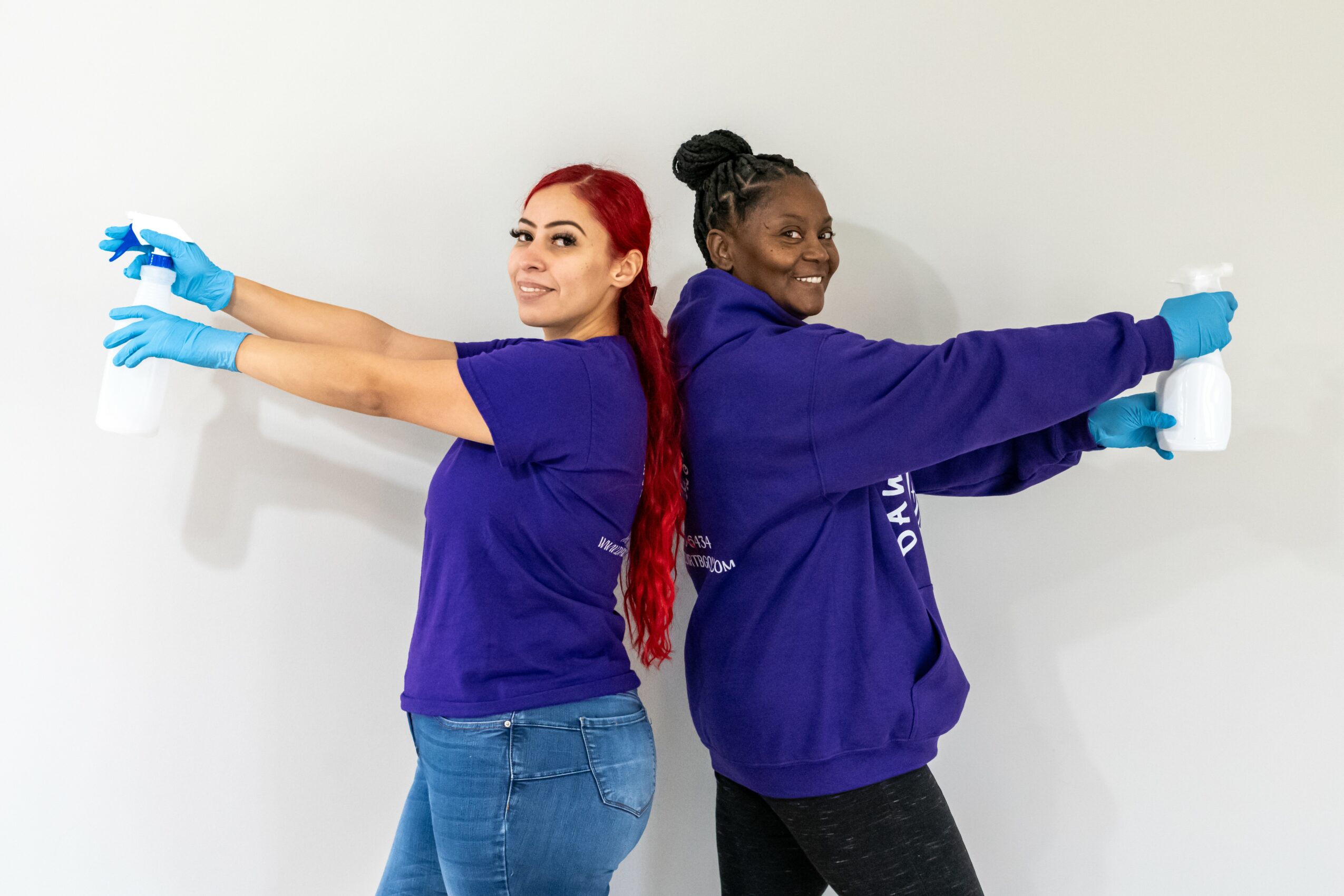Two women in blue shirts and gloves stand back-to-back, arms extended, smiling against a white wall.