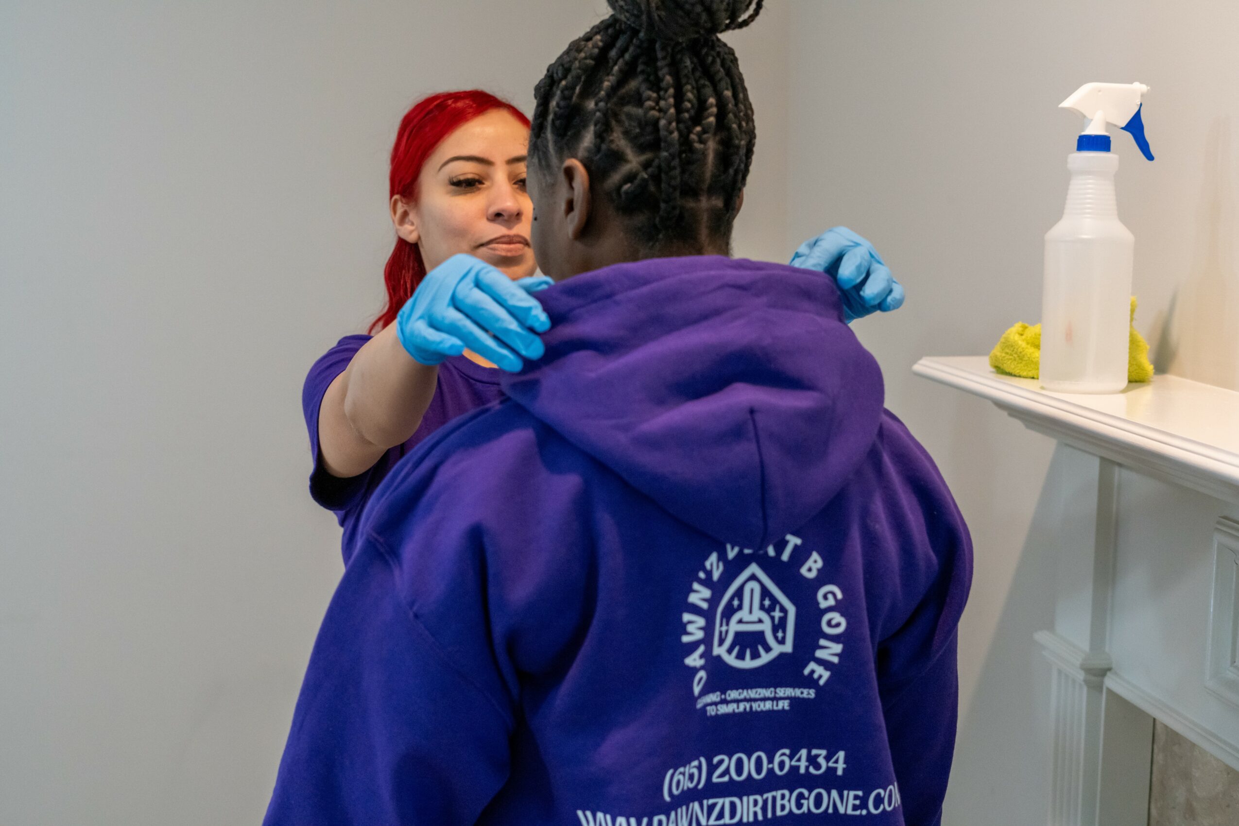 A healthcare worker with red hair and blue gloves examines a person wearing a purple hoodie in a clinical setting.