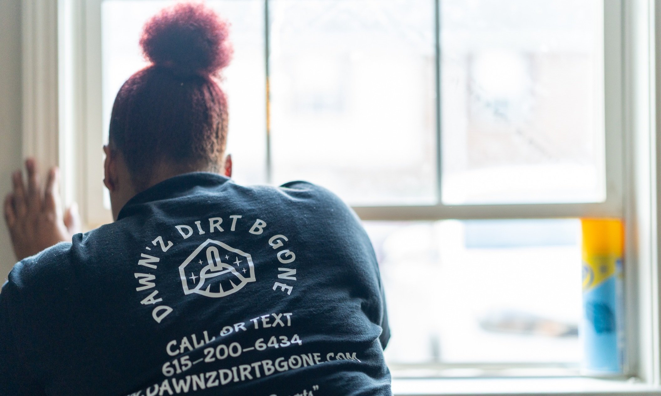 Person with red hair in a bun, wearing a dark shirt with white text and logo, sitting by a window, facing away.