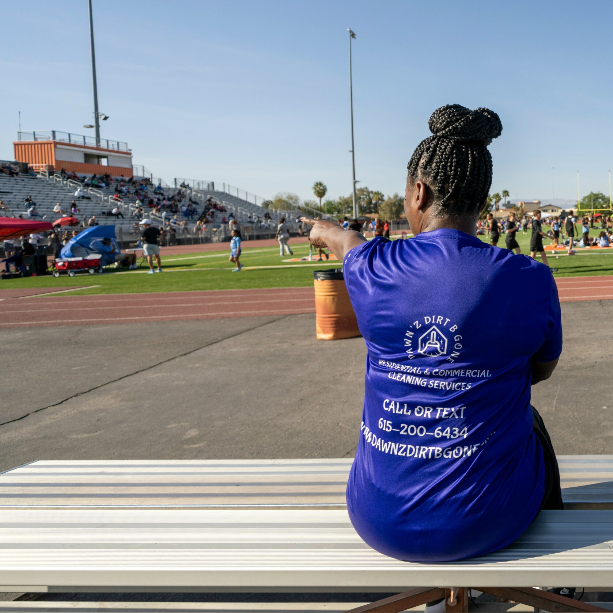 Woman with braided hair sitting on a bench at a track and field event, pointing towards the field, with spectators in the background.