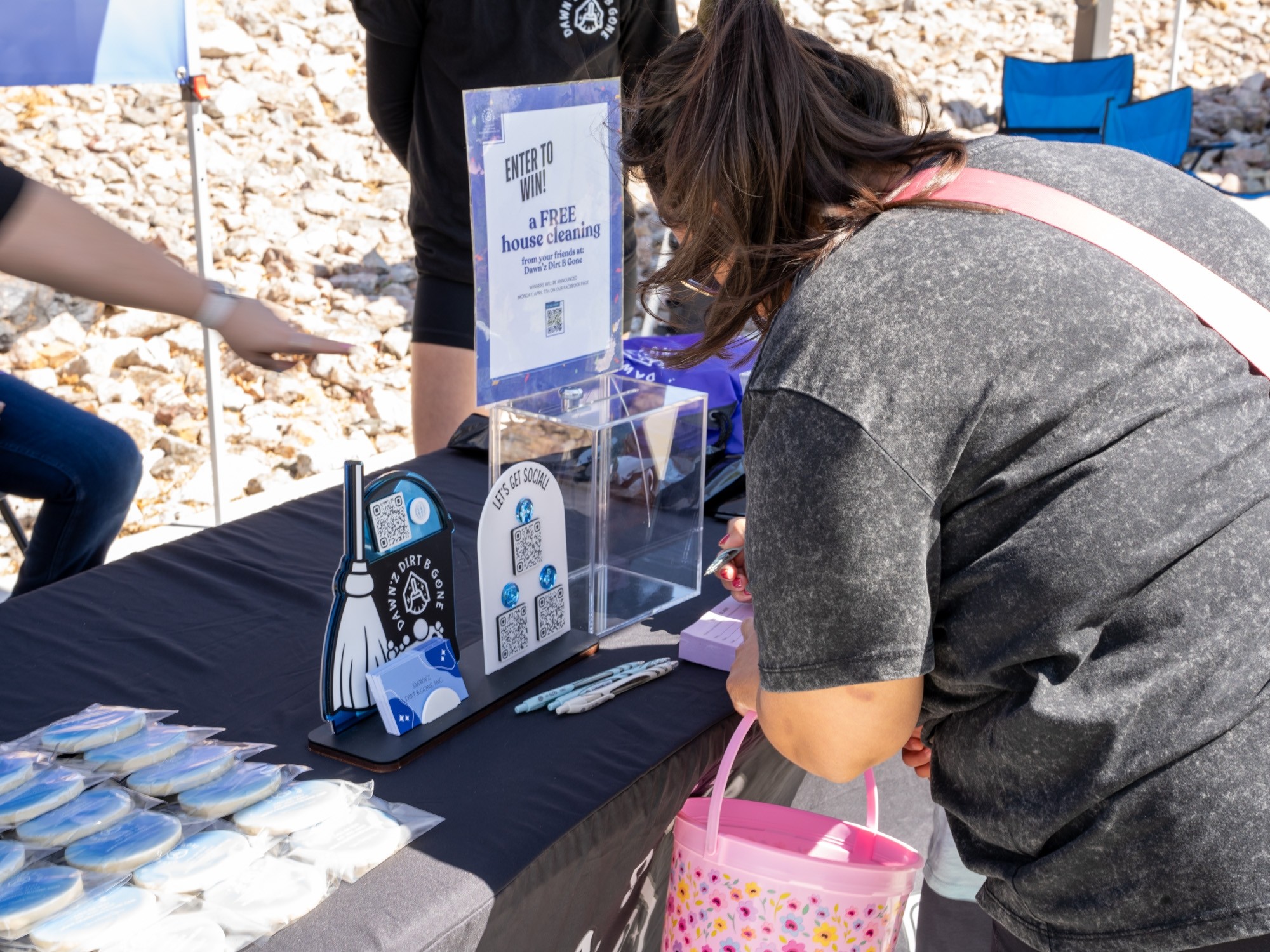 Person leaning over a table with informational displays and items, outdoors on rocky ground, with other people nearby.