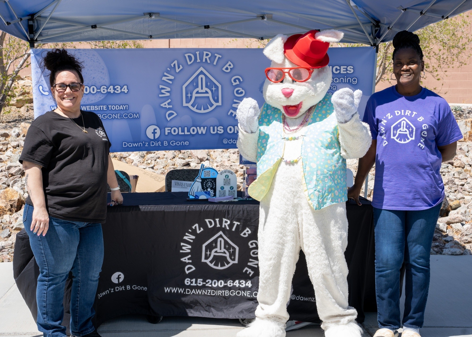Two women stand beside a person in a bunny costume at a booth with a blue banner, outdoor setting, sunny weather.