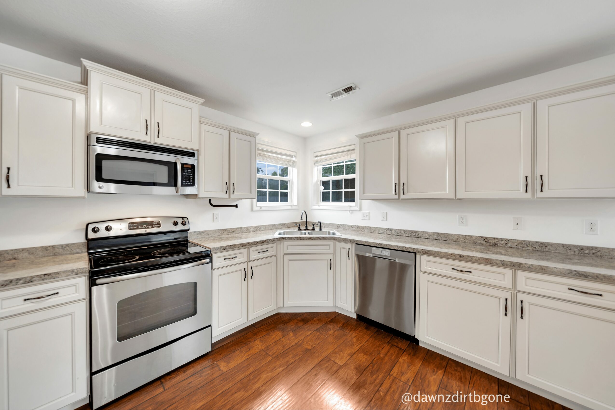 Kitchen with white cabinets, stainless steel appliances, and wooden floor, illuminated by natural light from windows.