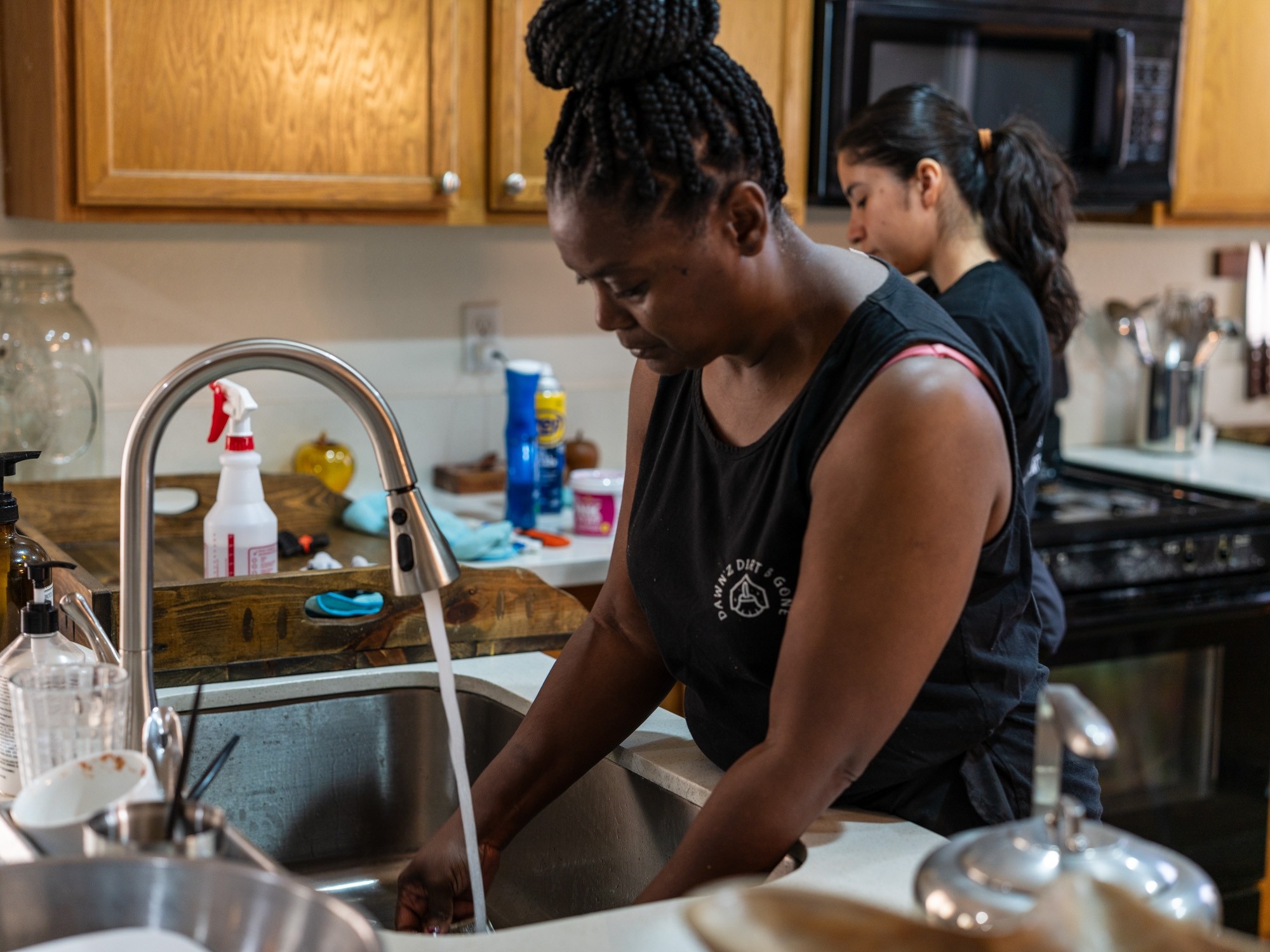 Two women washing dishes in a kitchen, one at the sink and the other in the background, with wooden cabinets and various kitchen items.