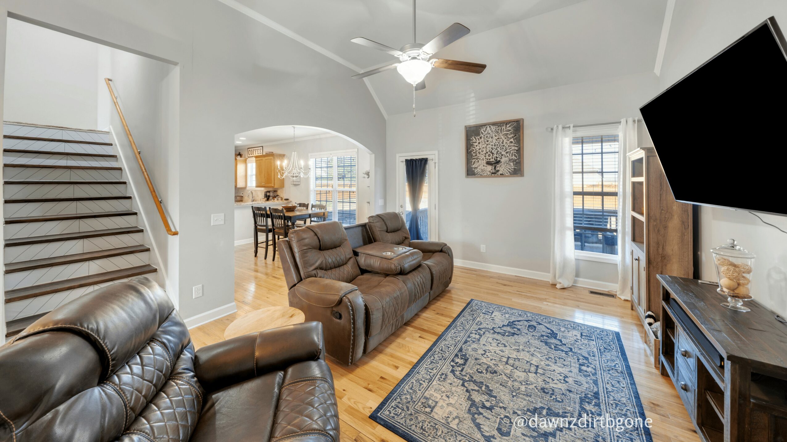 Living room with brown leather sofas, a blue patterned rug, a wall-mounted TV, and large windows with curtains.