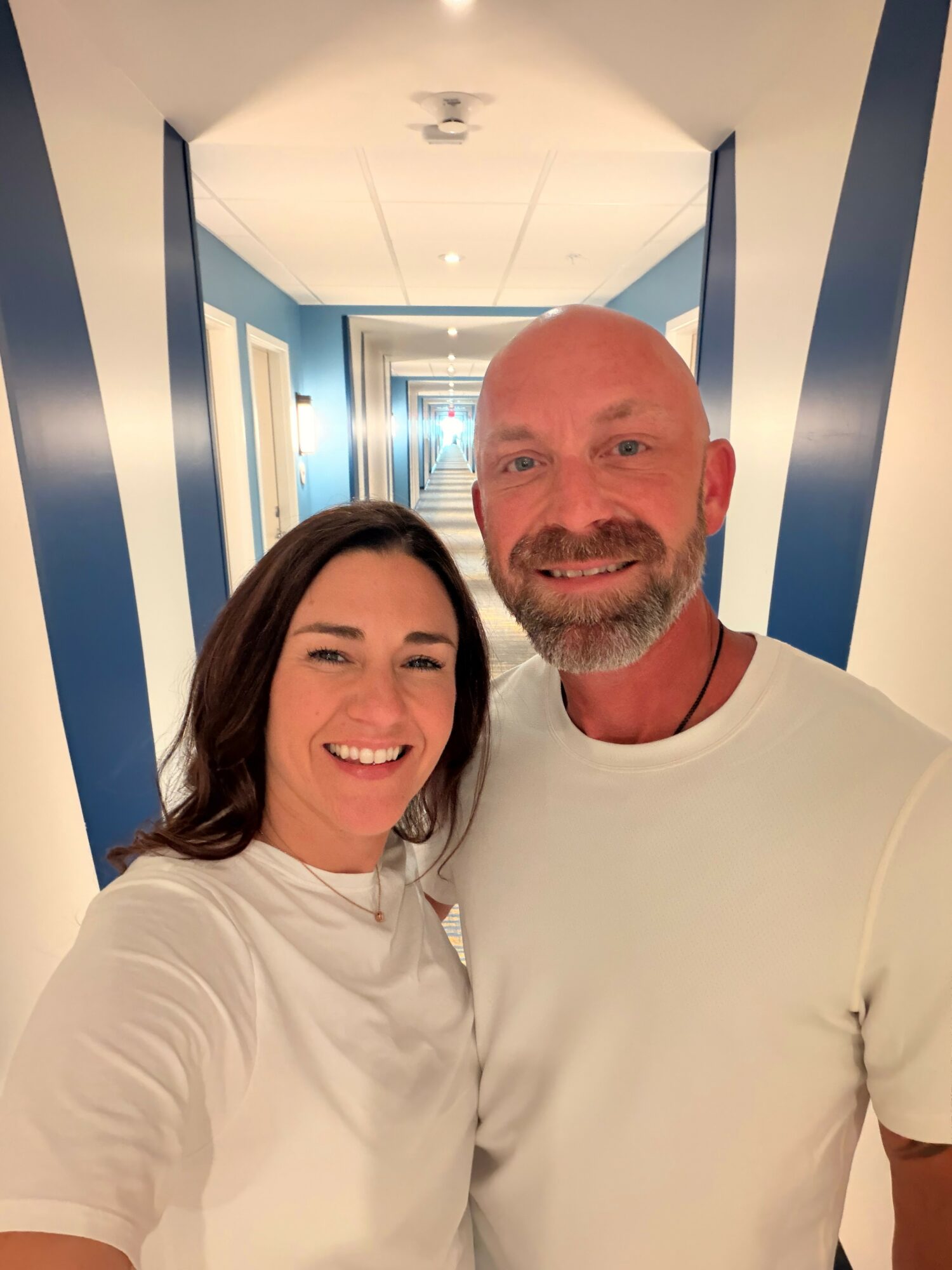 Two smiling people, a woman with dark hair and a man with a beard, stand in a brightly lit hallway with blue walls.
