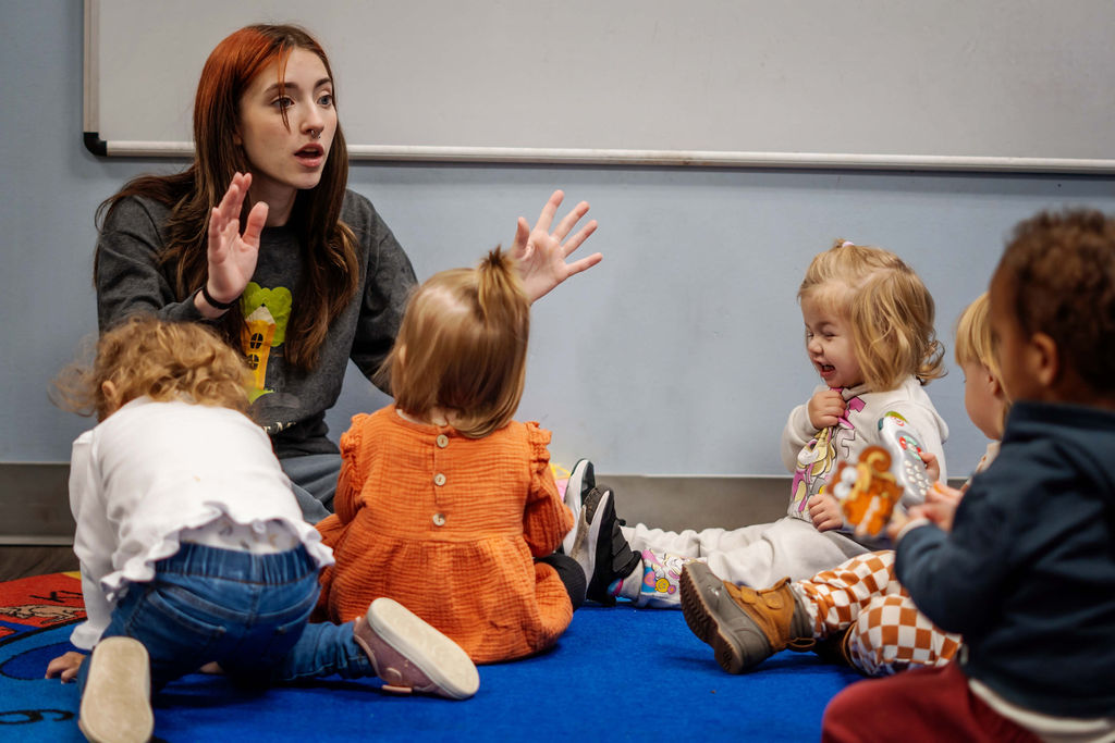 Young woman speaking to a group of children sitting on a blue carpet in a classroom, whiteboard in background.