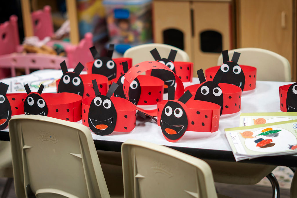 Table with red and black ladybug-themed paper crafts and children's drawings, surrounded by white chairs in a classroom or playroom.