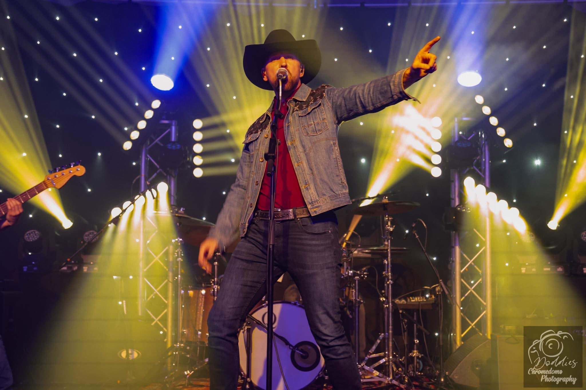 Performer wearing a cowboy hat, denim jacket, and jeans on stage with colorful lights and musical equipment.