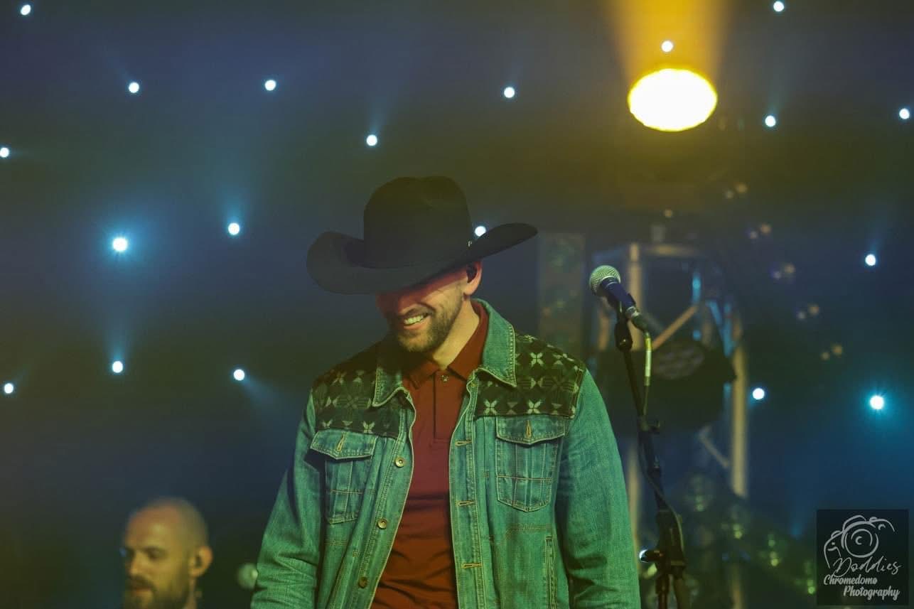 Person wearing a large hat, denim jacket, and red shirt, smiling on stage with starry background.