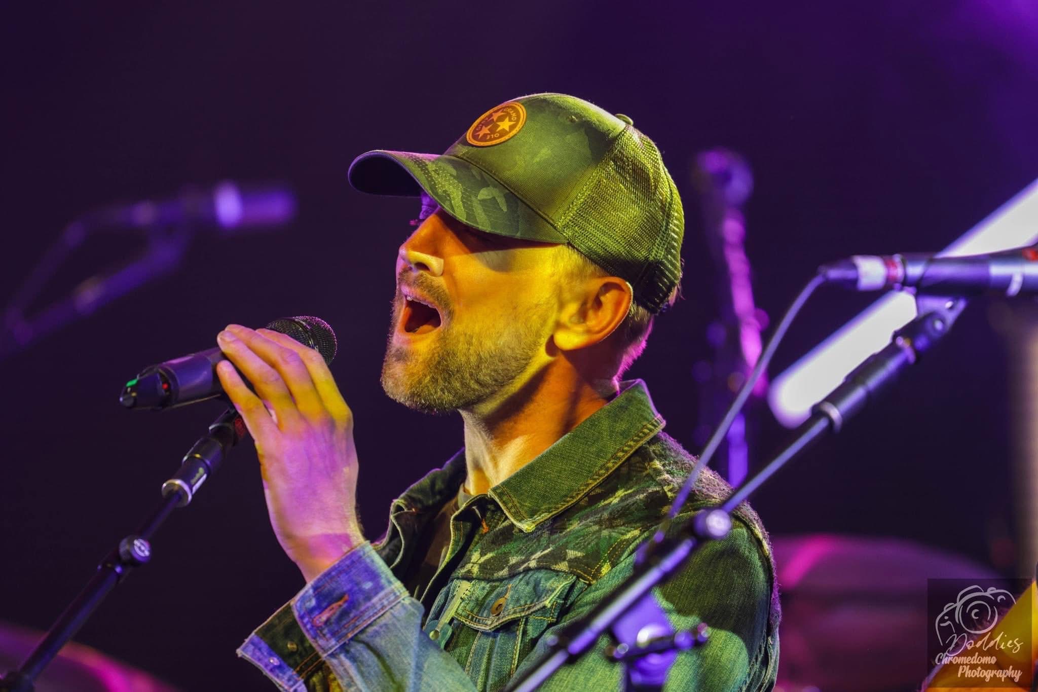 Man singing into a microphone on stage, wearing a cap and camouflage jacket, with purple stage lighting.