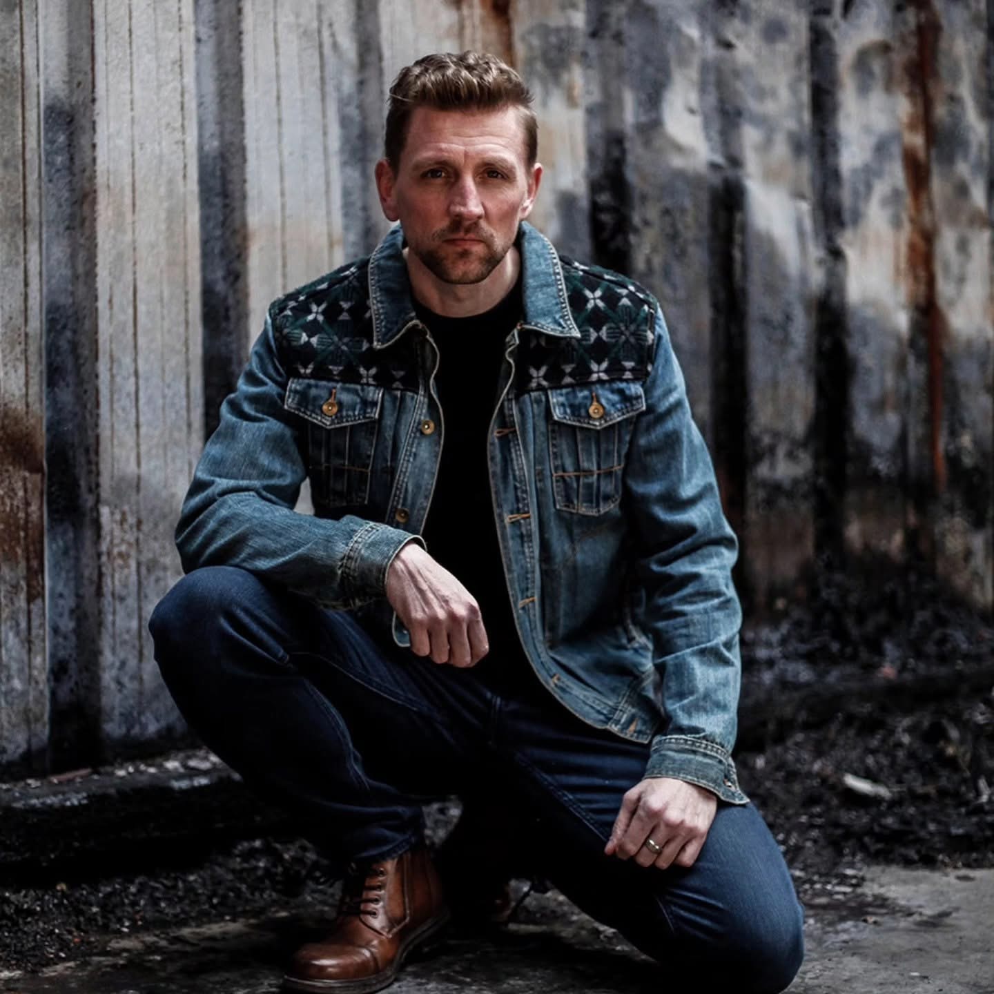 Man crouching in front of a weathered wooden fence, wearing a denim jacket and jeans, with a serious expression.