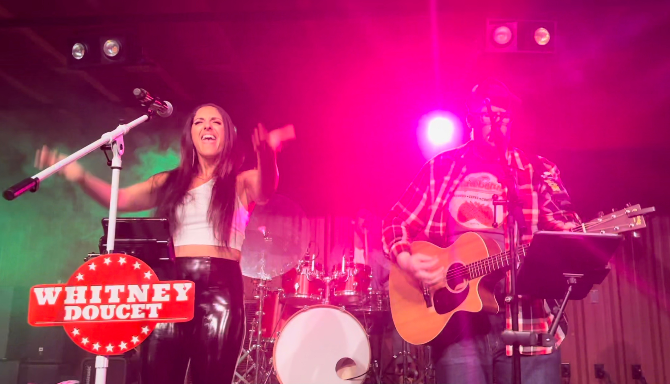 Two performers on stage, one singing and the other playing guitar, with colorful lighting and a Whitney Doucet sign.
