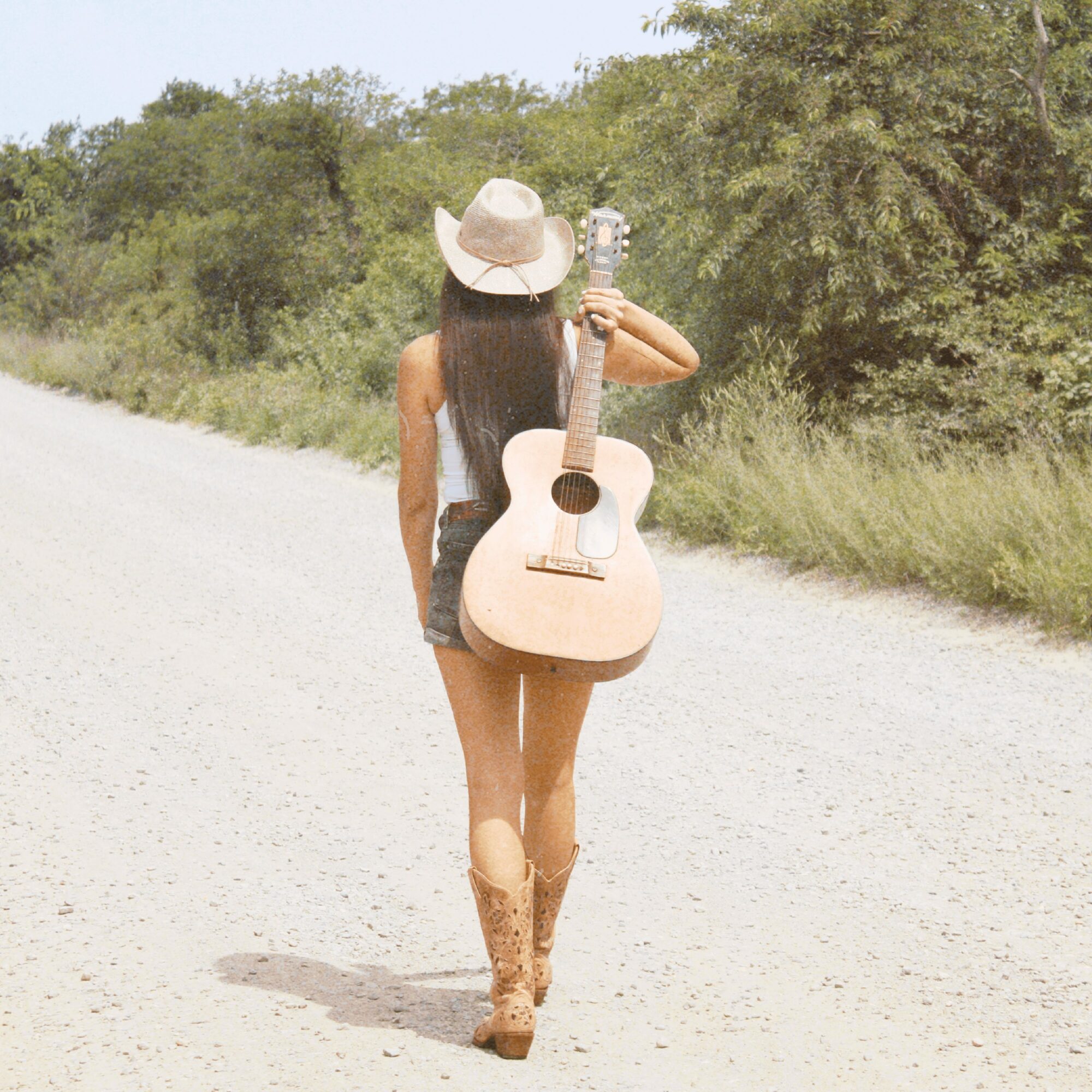 Person walking on a dirt road holding a guitar, wearing a hat and cowboy boots, with trees in the background.