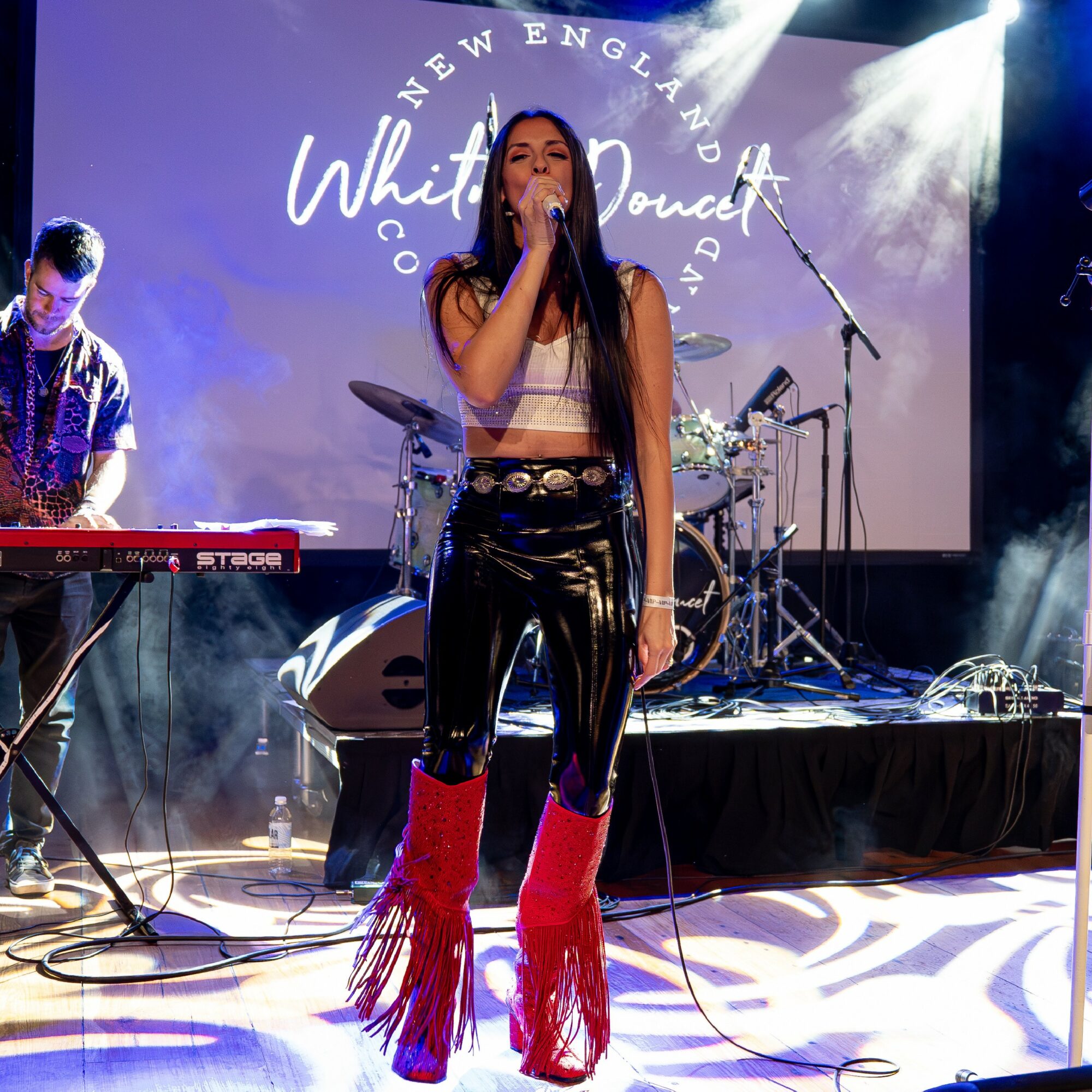 Female singer performing on stage with band, microphone in hand, wearing shiny black pants and red fringed boots, stage lights shining.