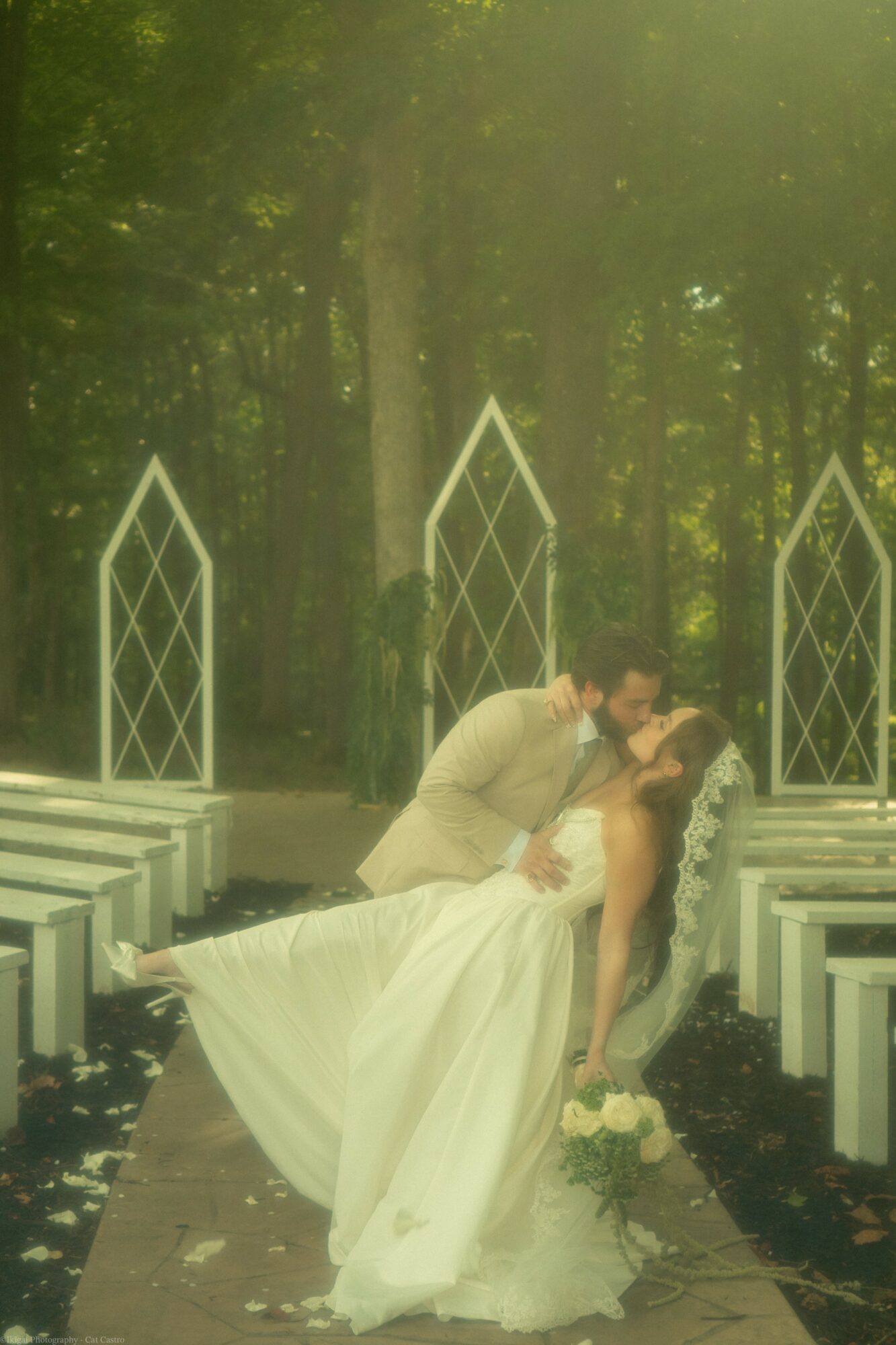 A couple in wedding attire dancing outdoors among white benches and tall trees, with sunlight filtering through leaves.